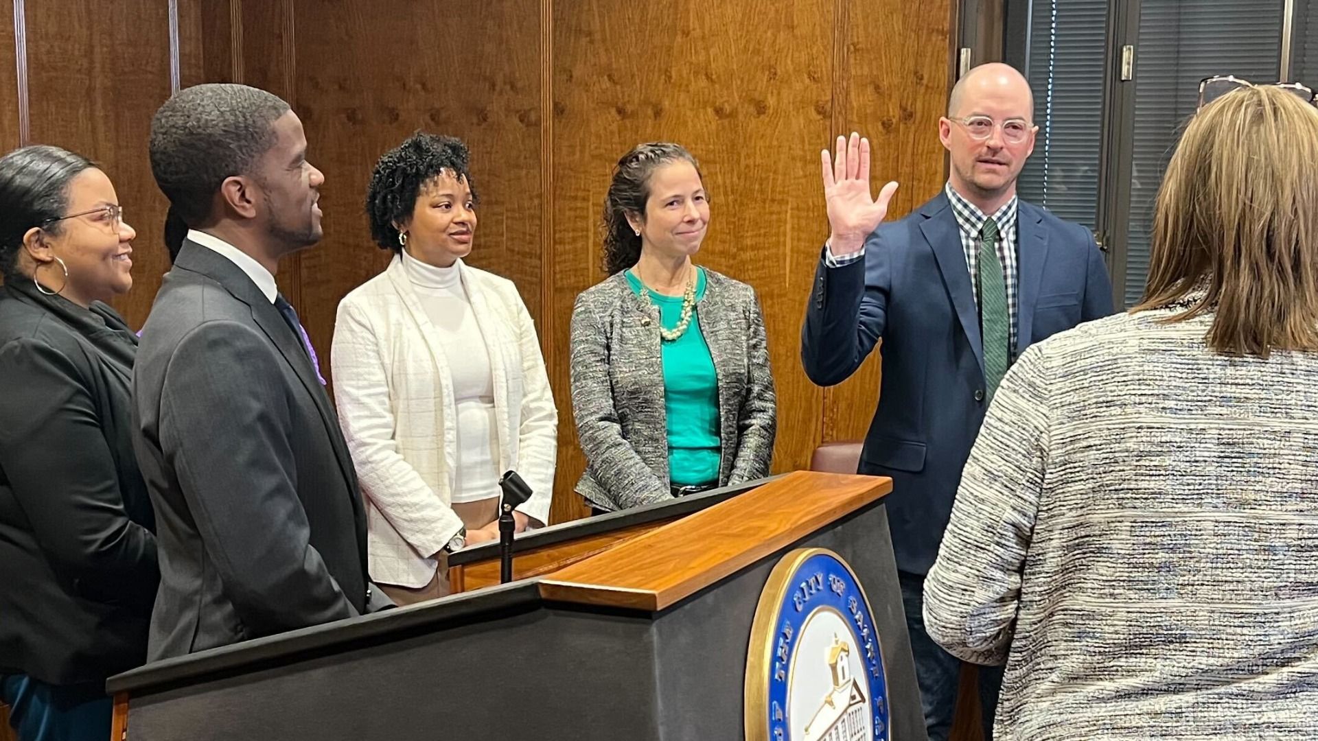A man in a suit stands with his hand raised and a group of other professionally-dressed people behind a lectern with the City of St. Paul logo on it