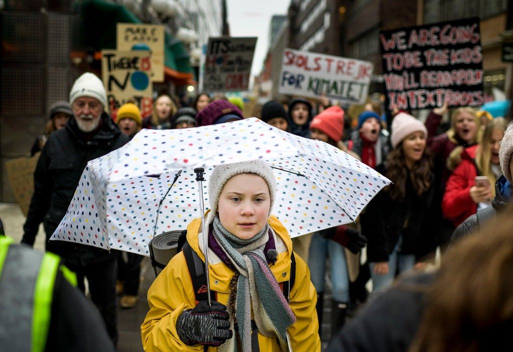Greta Thunberg walks down the street with other protestors, carrying an umbrella. 