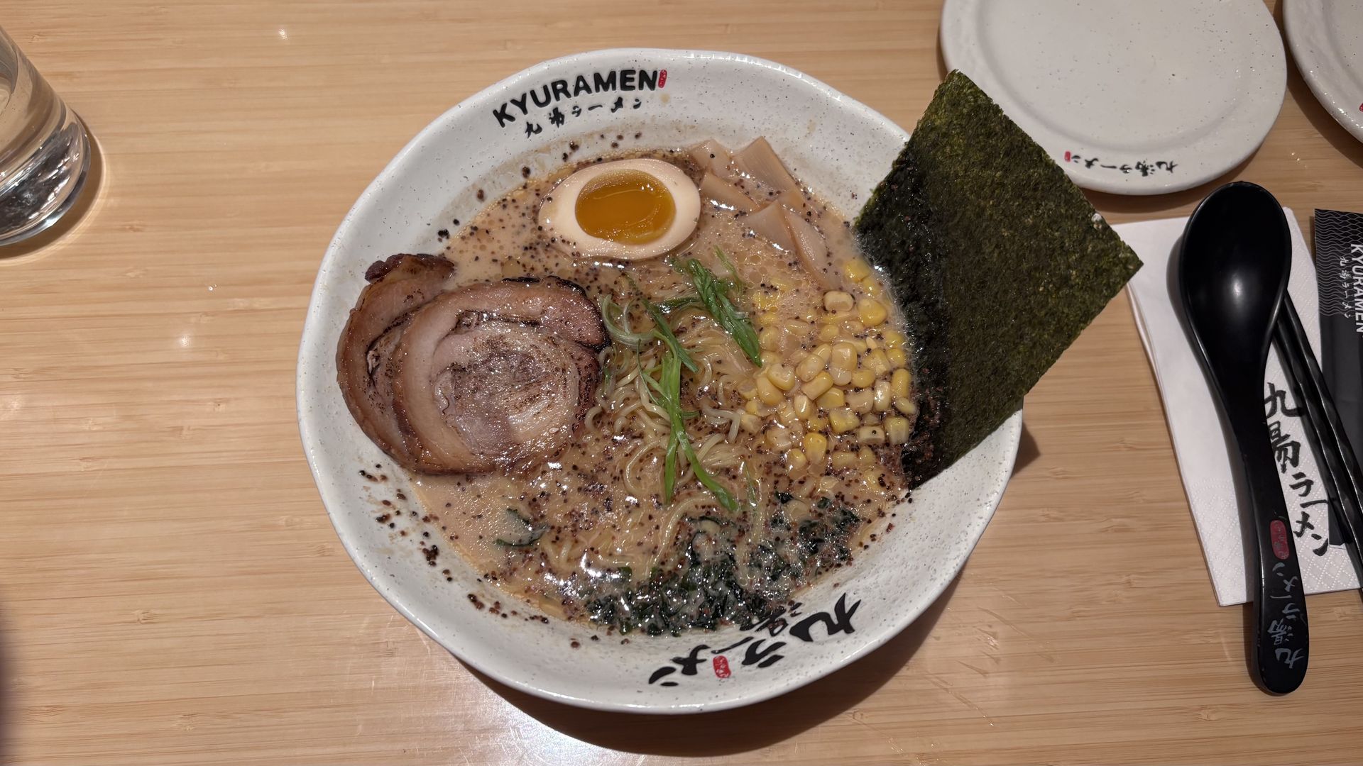 Ramen in a white speckled bowl topped with a large chashu pork slice, a halved soft-boiled egg, corn, nori, green onions, and bamboo shoots in a creamy broth; wooden table and utensils nearby.