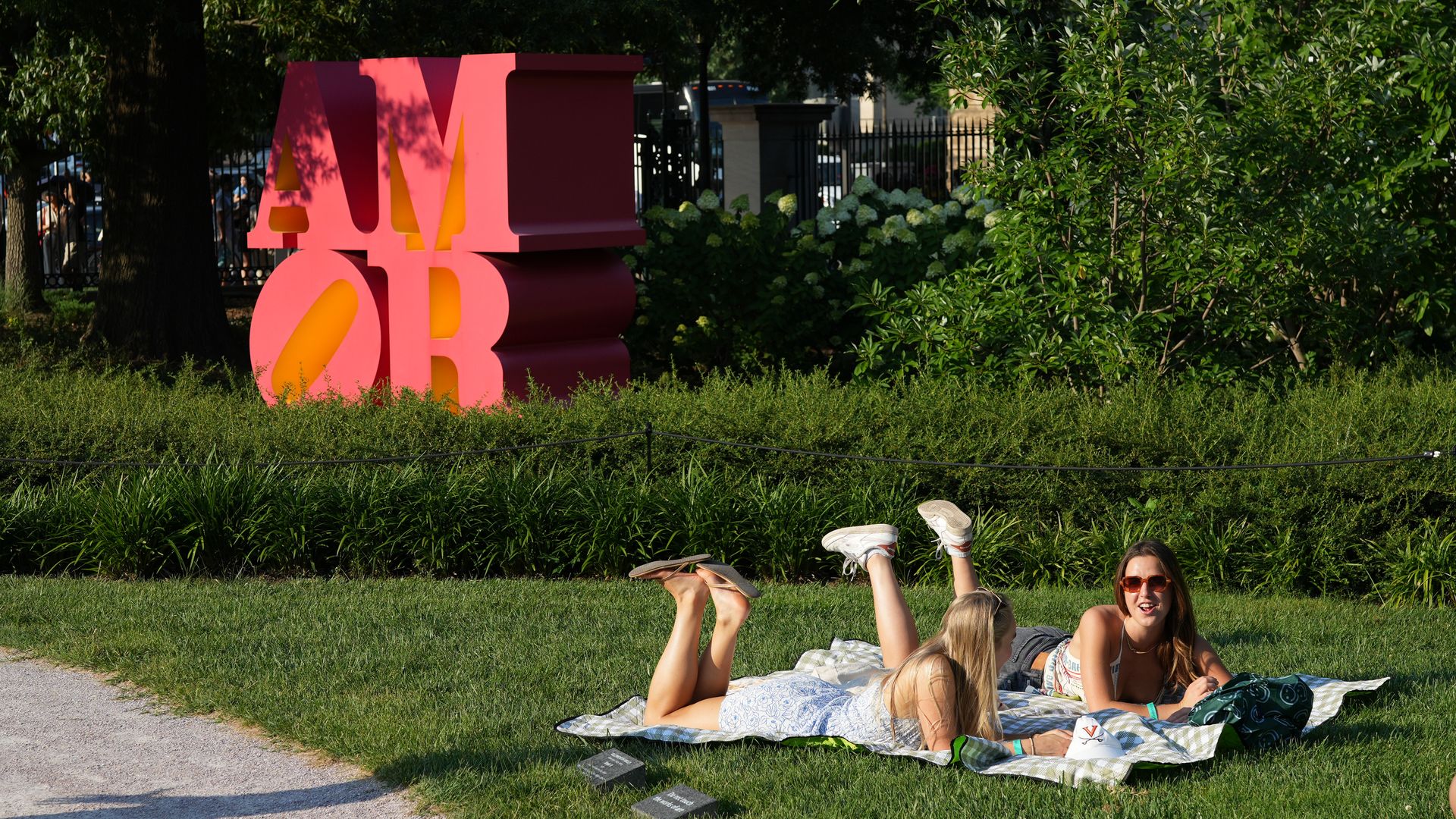 Bright pink sculpture spelling AMOR with orange highlights sits in a sunny park, behind green trees; a group of friends lounges on a blanket in the grass foreground.