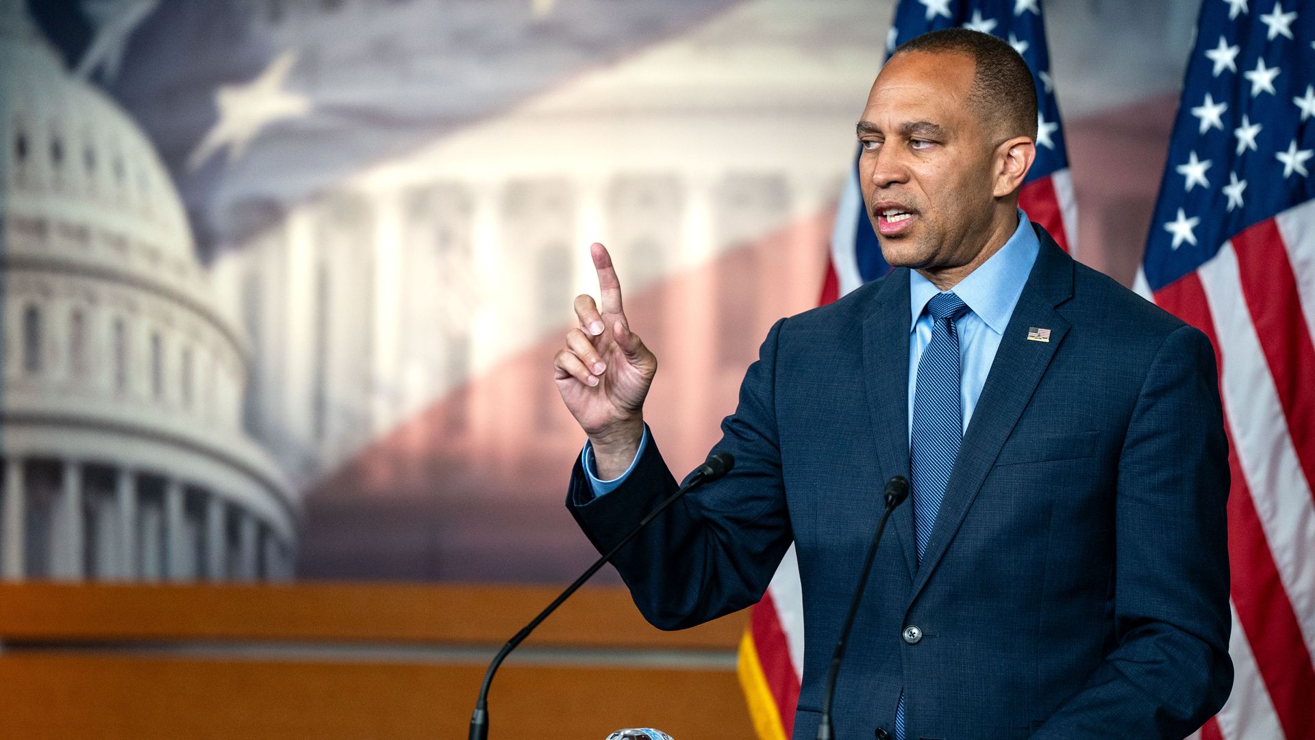 House Minority Leader Hakeem Jeffries, wearing a blue suit and speaking in front of a Capitol mural and American flags.