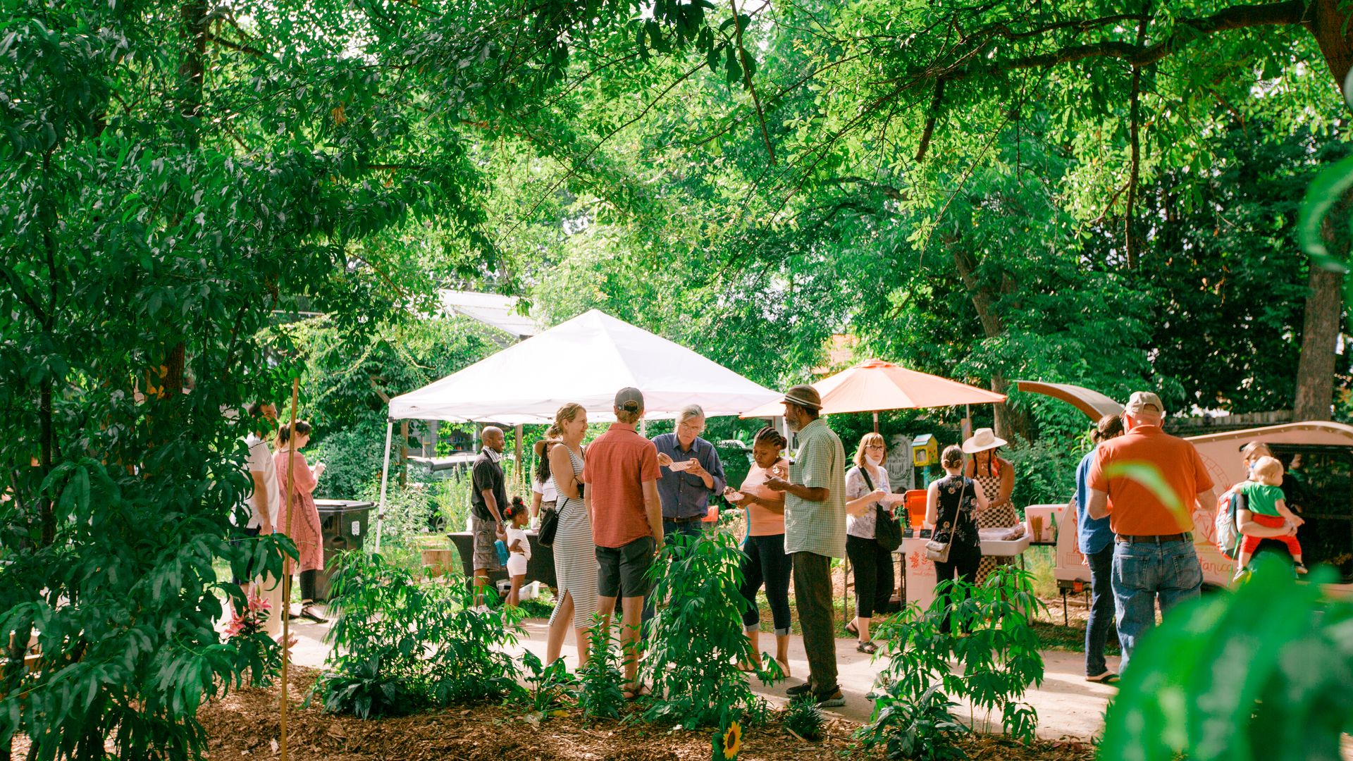 People talk, sample food, and peruse vendor stands at a lush green farmers market in East Atlanta Village