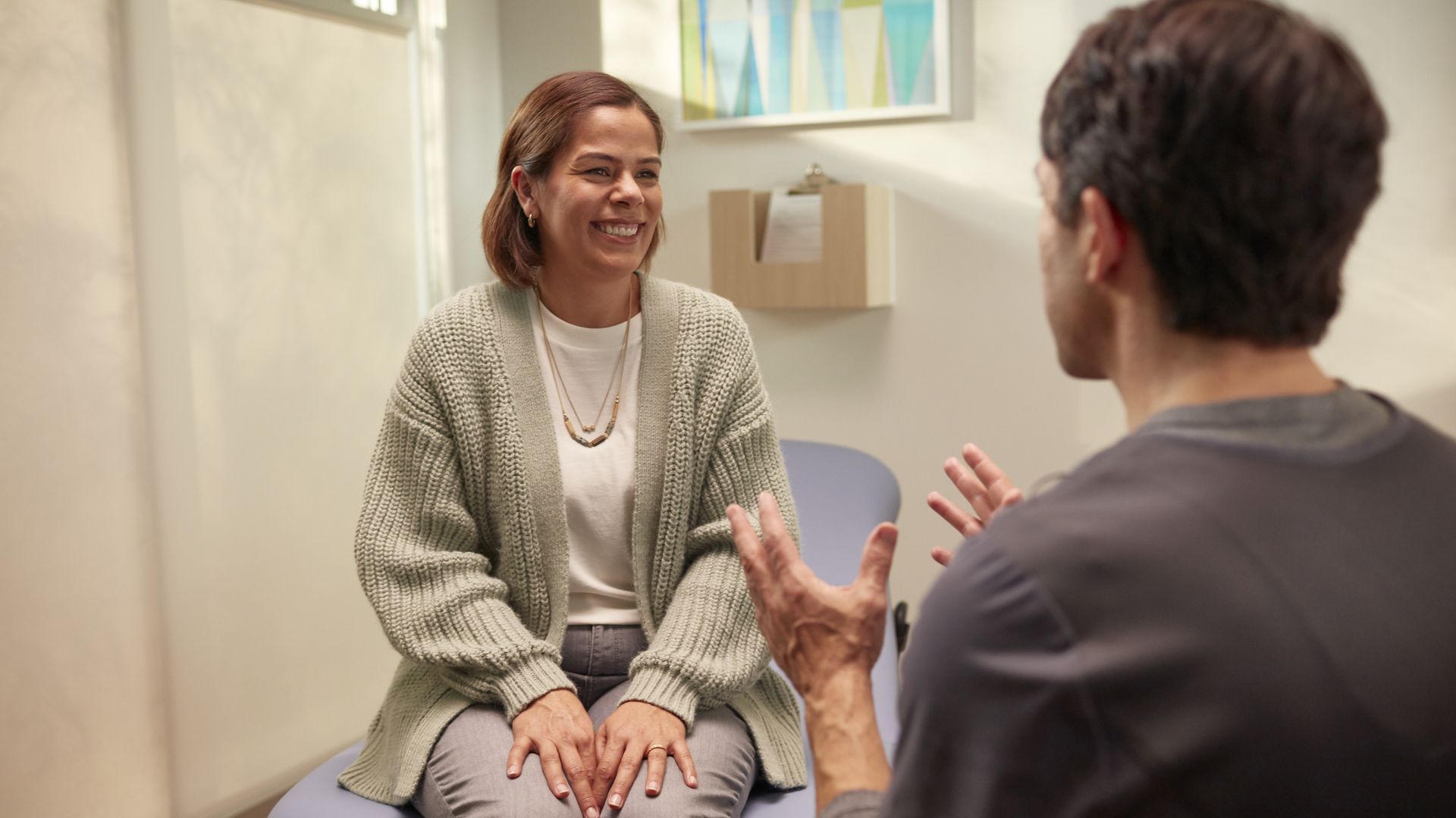 A woman in a light gray cardigan sits on a blue exam chair, smiling while talking to a man with his back to the camera; they're in a bright room with geometric wall art behind them.