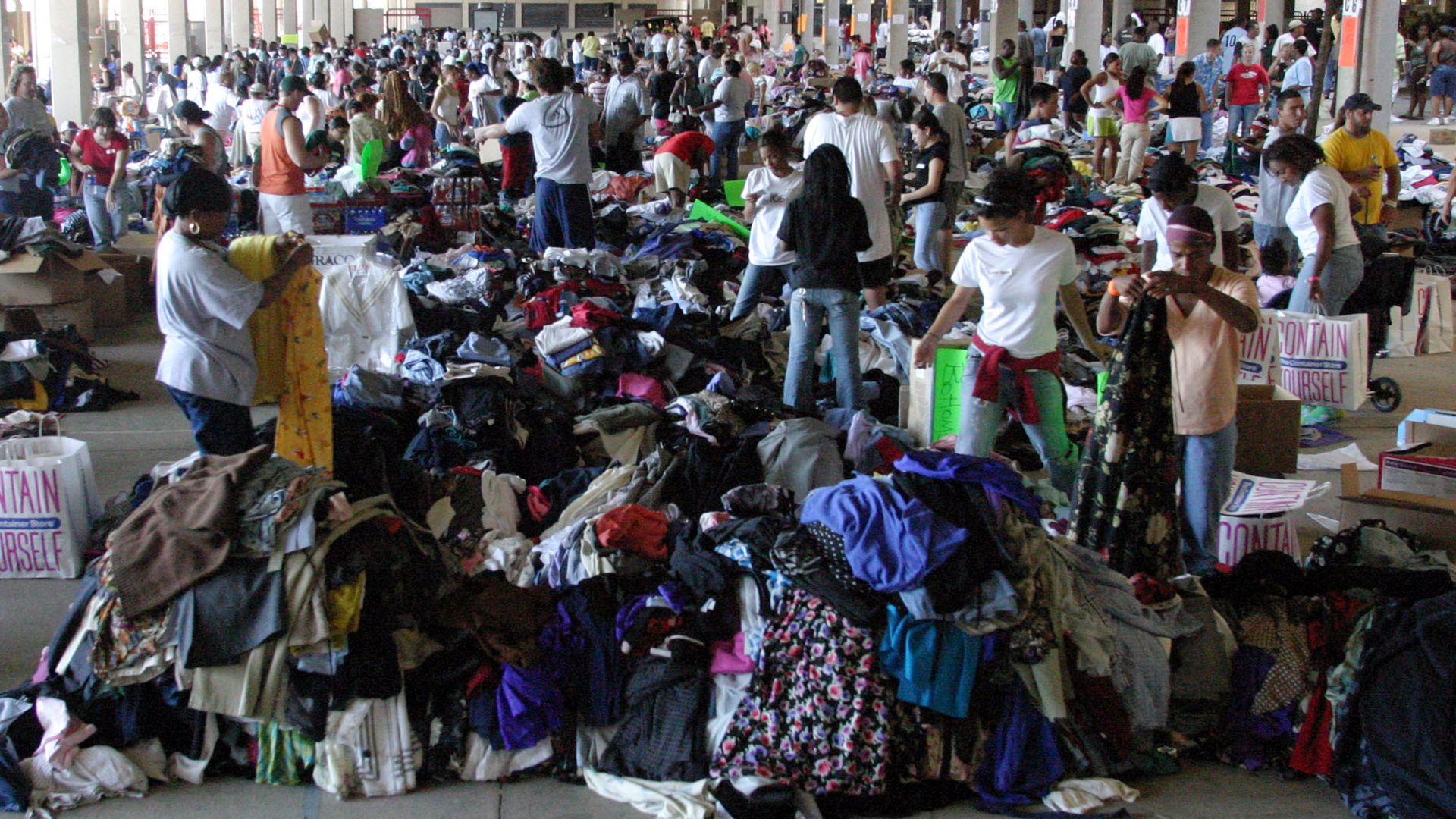 Large indoor space filled with people sorting through numerous piles of donated clothes in various colors, with boxes and bags scattered around.