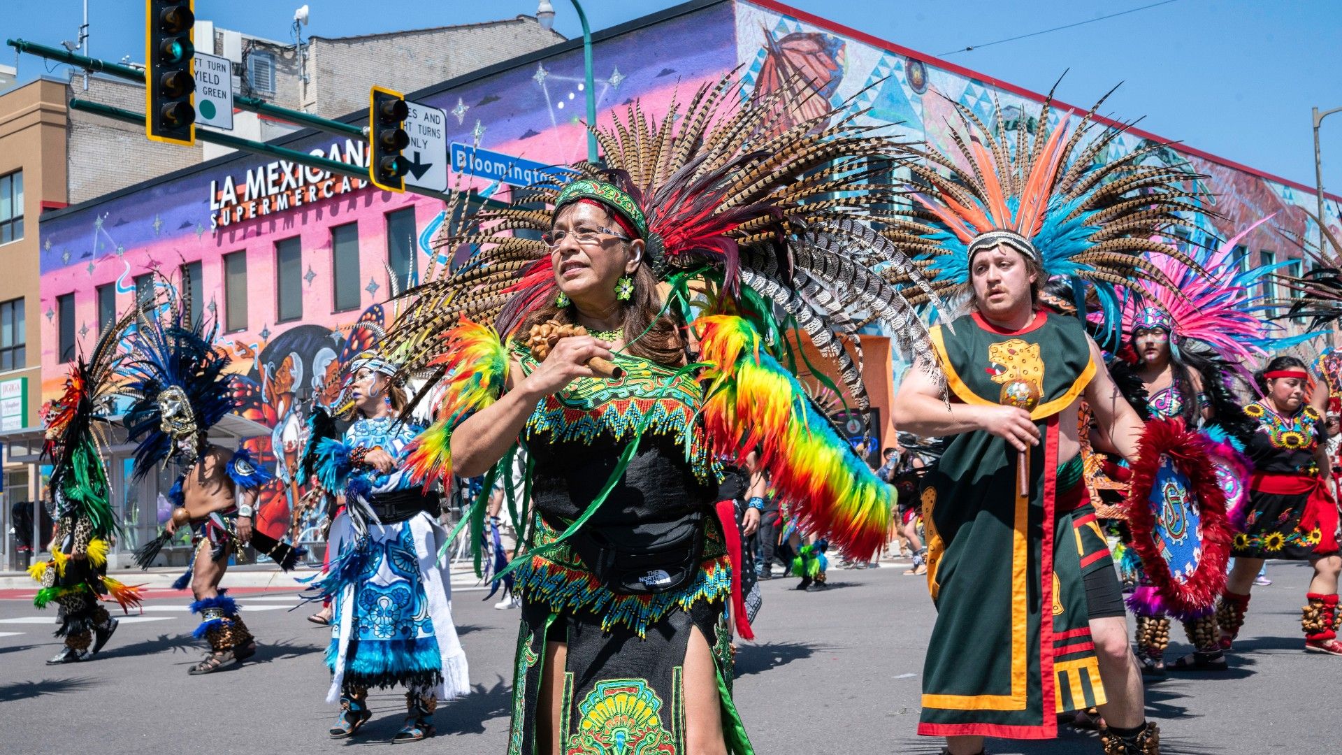 Performers in colorful traditional attire dance during a lively cultural festival on Lake Street, with vibrant murals and a local market visible in the background.