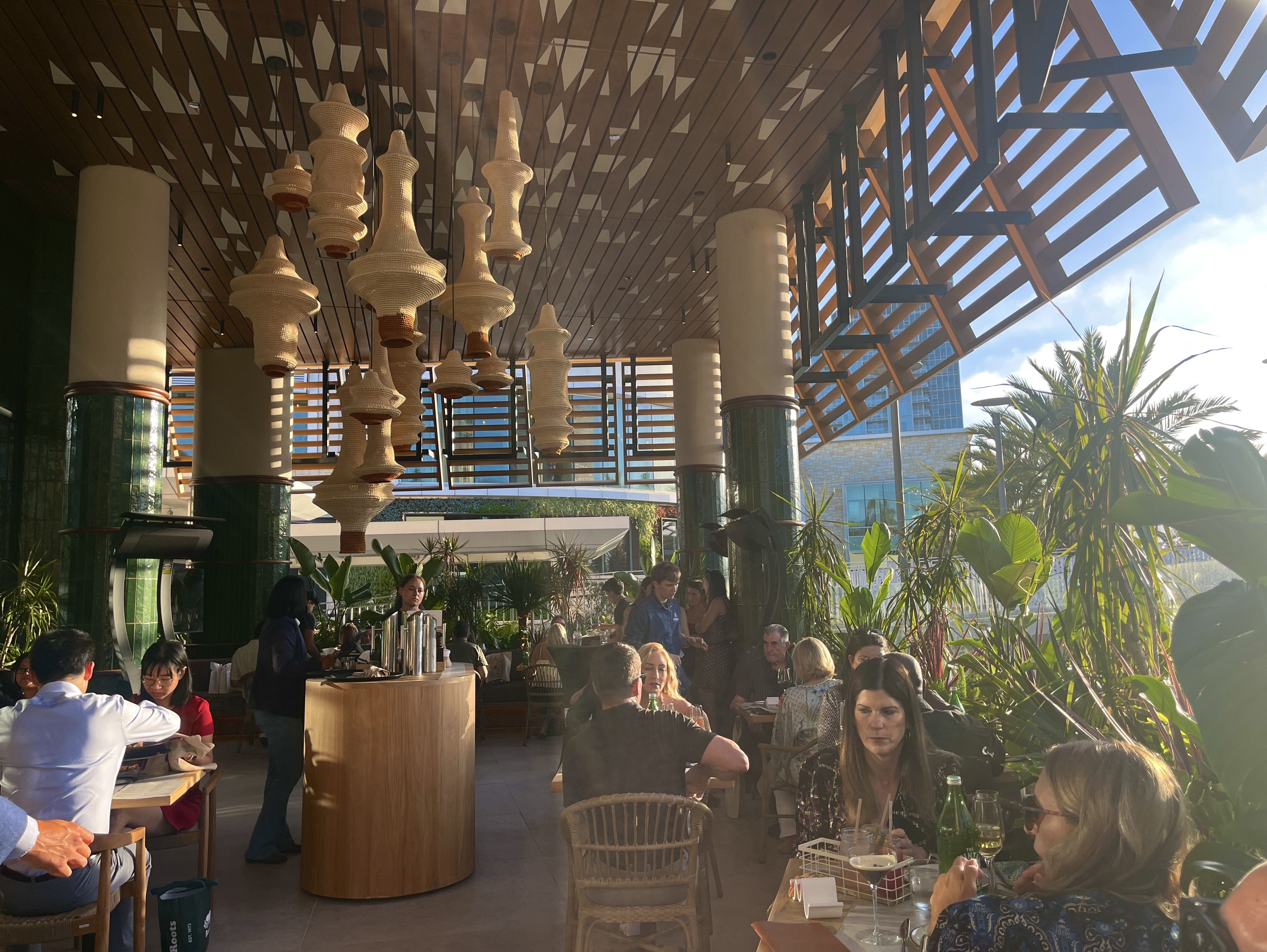 Restaurant patio with wicker chandeliers hanging from a wooden ceiling; diners at tables among palms, with a blue sky visible through a lattice structure.