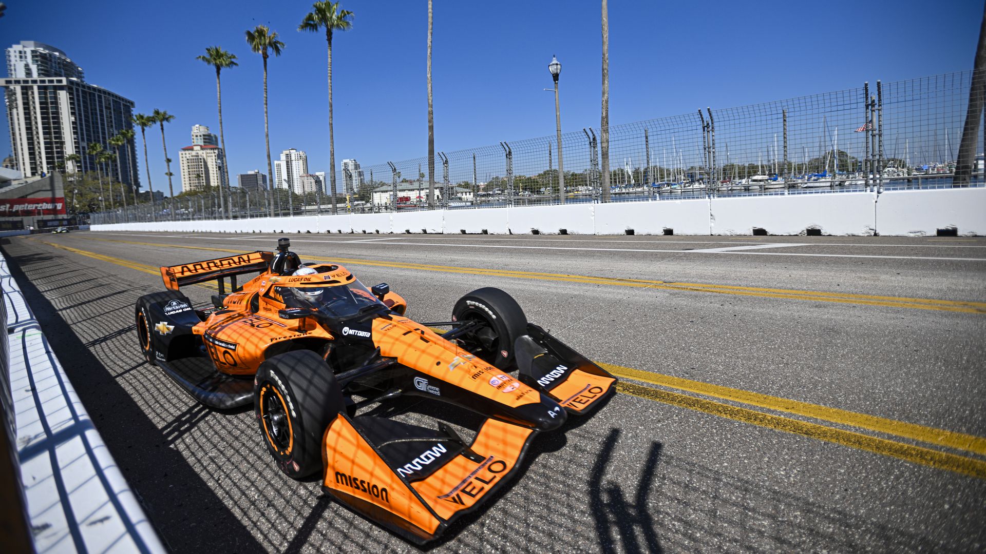 Orange and black IndyCar race car on a street circuit with palm trees and high-rise buildings under a clear blue sky in St. Petersburg.