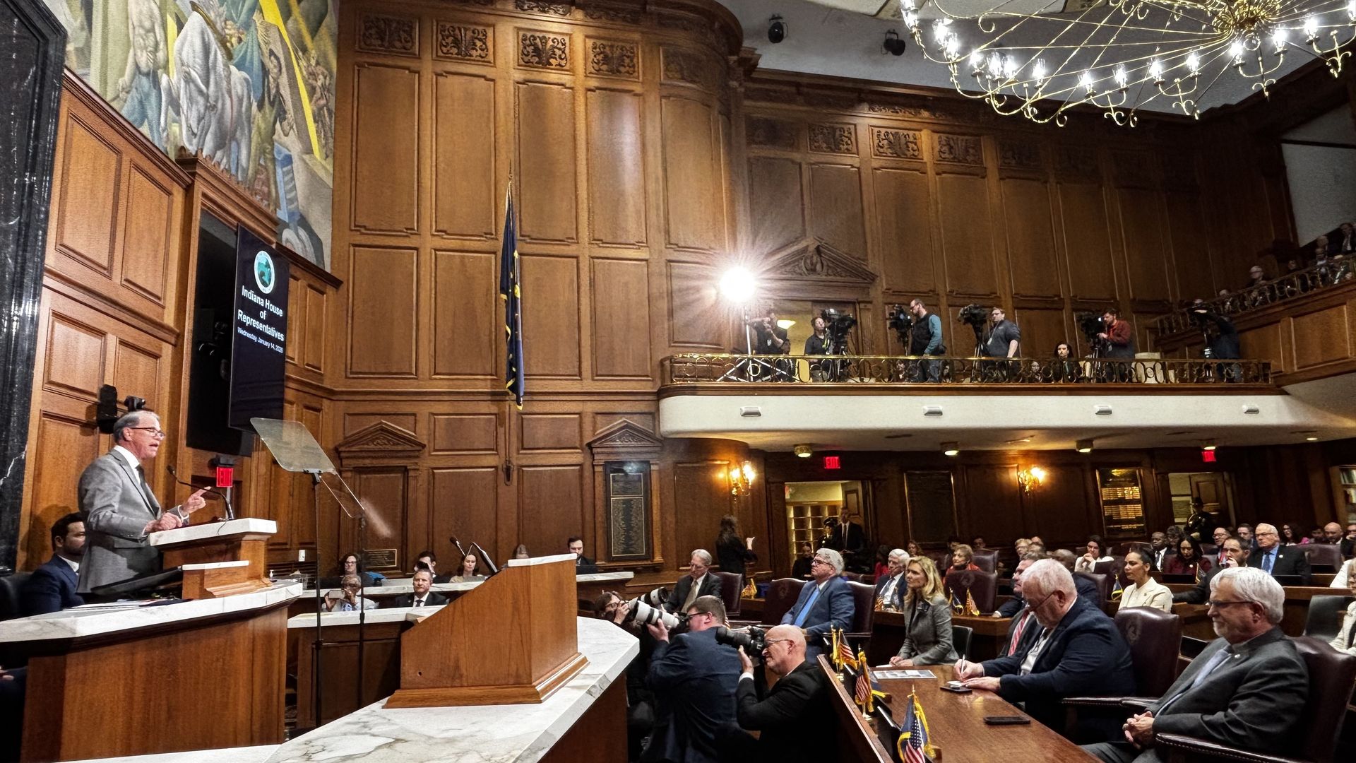 Speaker in gray suit at podium addressing Indiana House of Representatives chamber with wood paneling, photographers, seated audience, and large chandelier overhead.