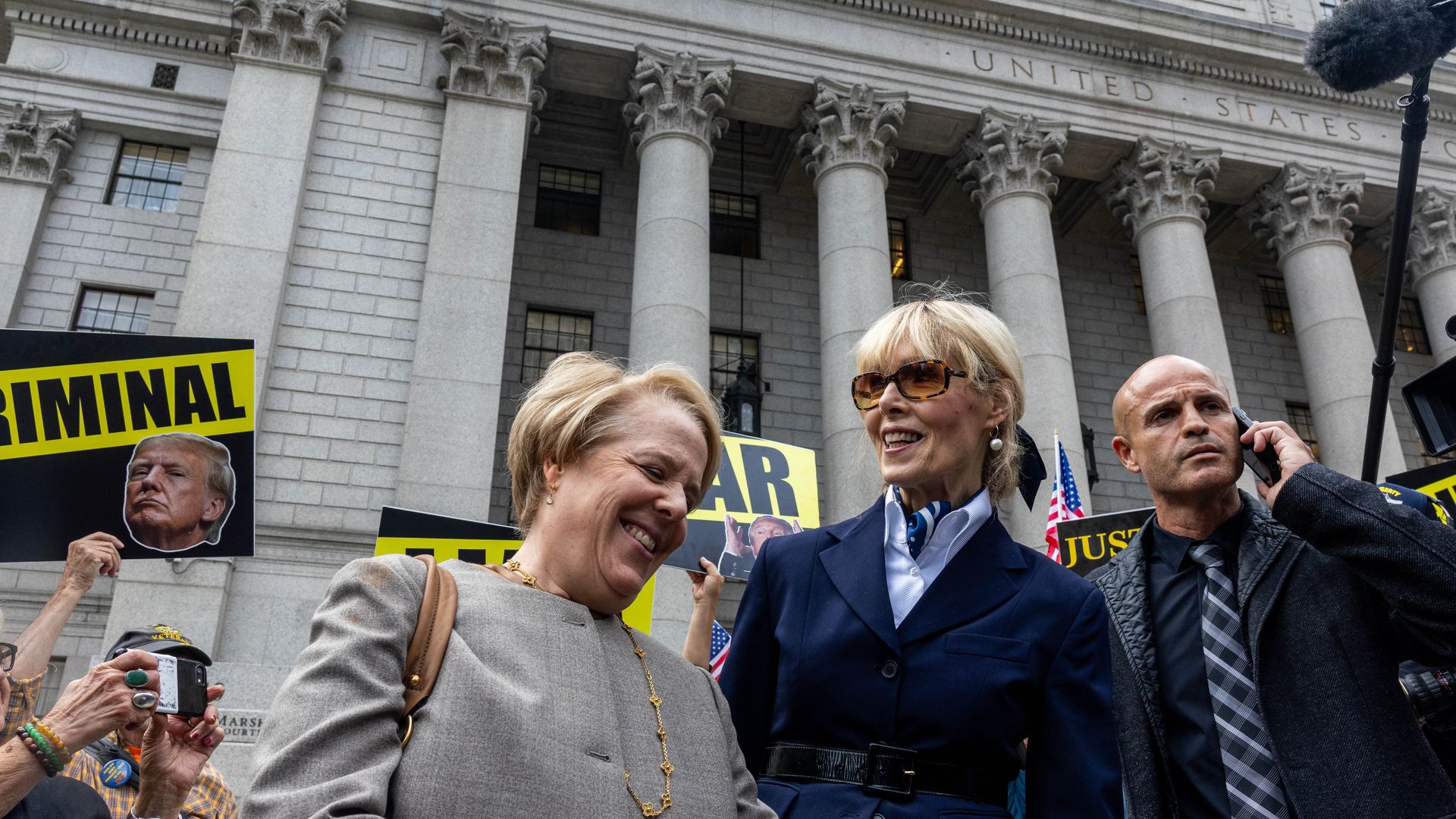 Two blonde women smile as they leave a courthouse as a crowd holds signs around them with Donald Trump's face on them.