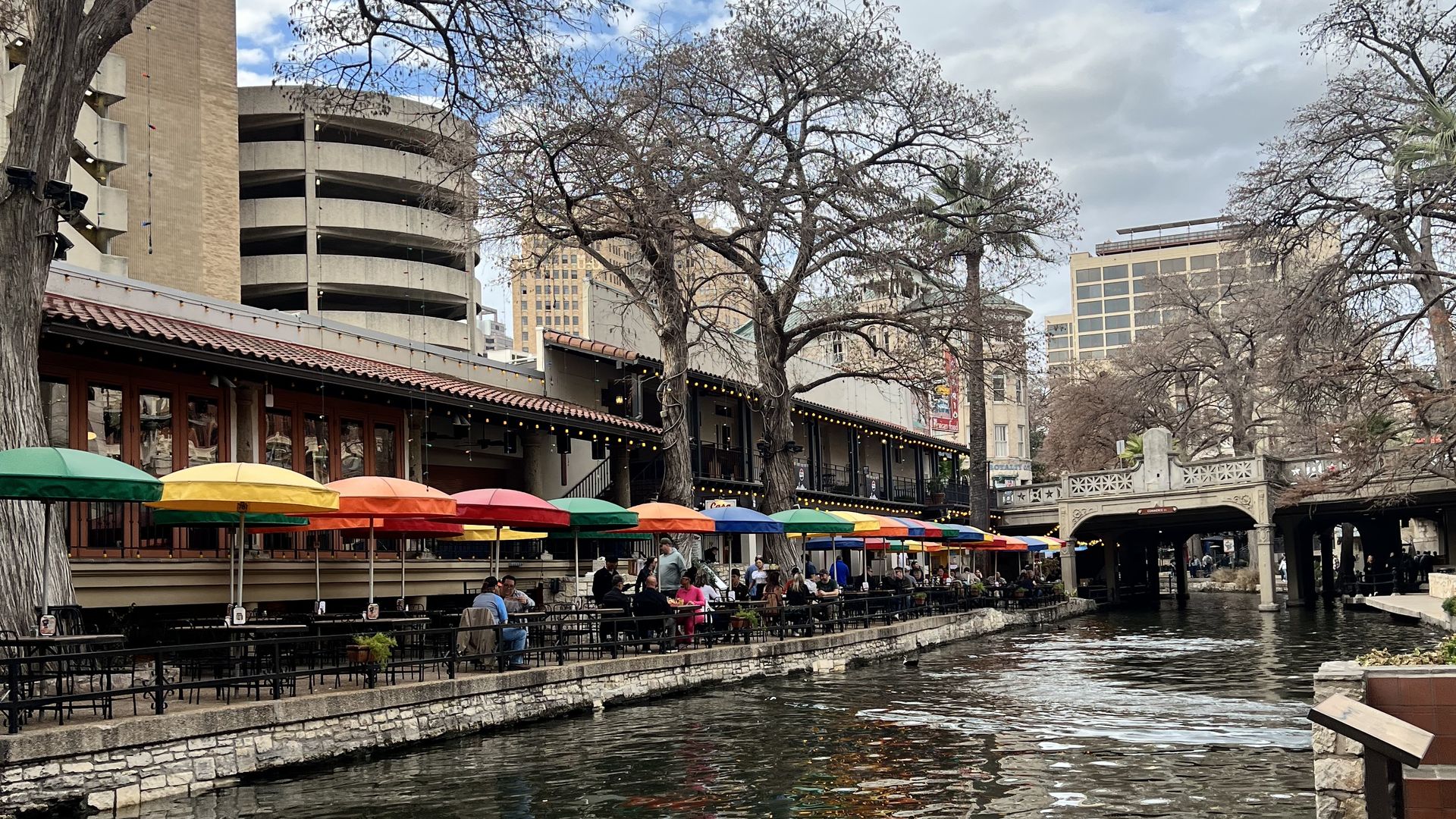 Riverside cafe row with colorful umbrellas along a stone canal wall; people dine under leafless trees while city buildings and a cloudy blue sky loom above, water shimmering with reflections.