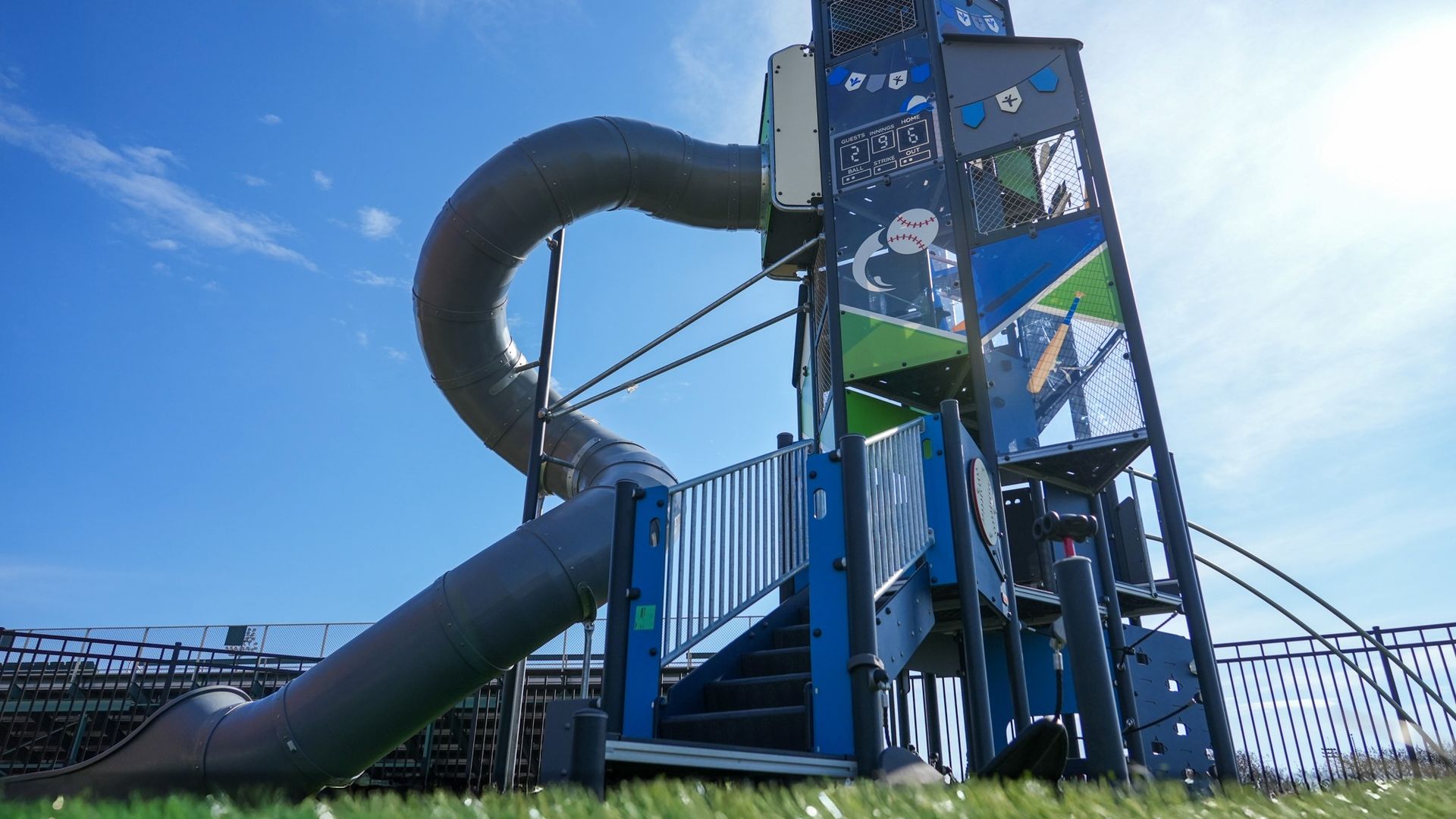 Blue and green playground slide structure with baseball-themed decorations and a spiral metal tube slide, set against a clear blue sky and green grass in the foreground.