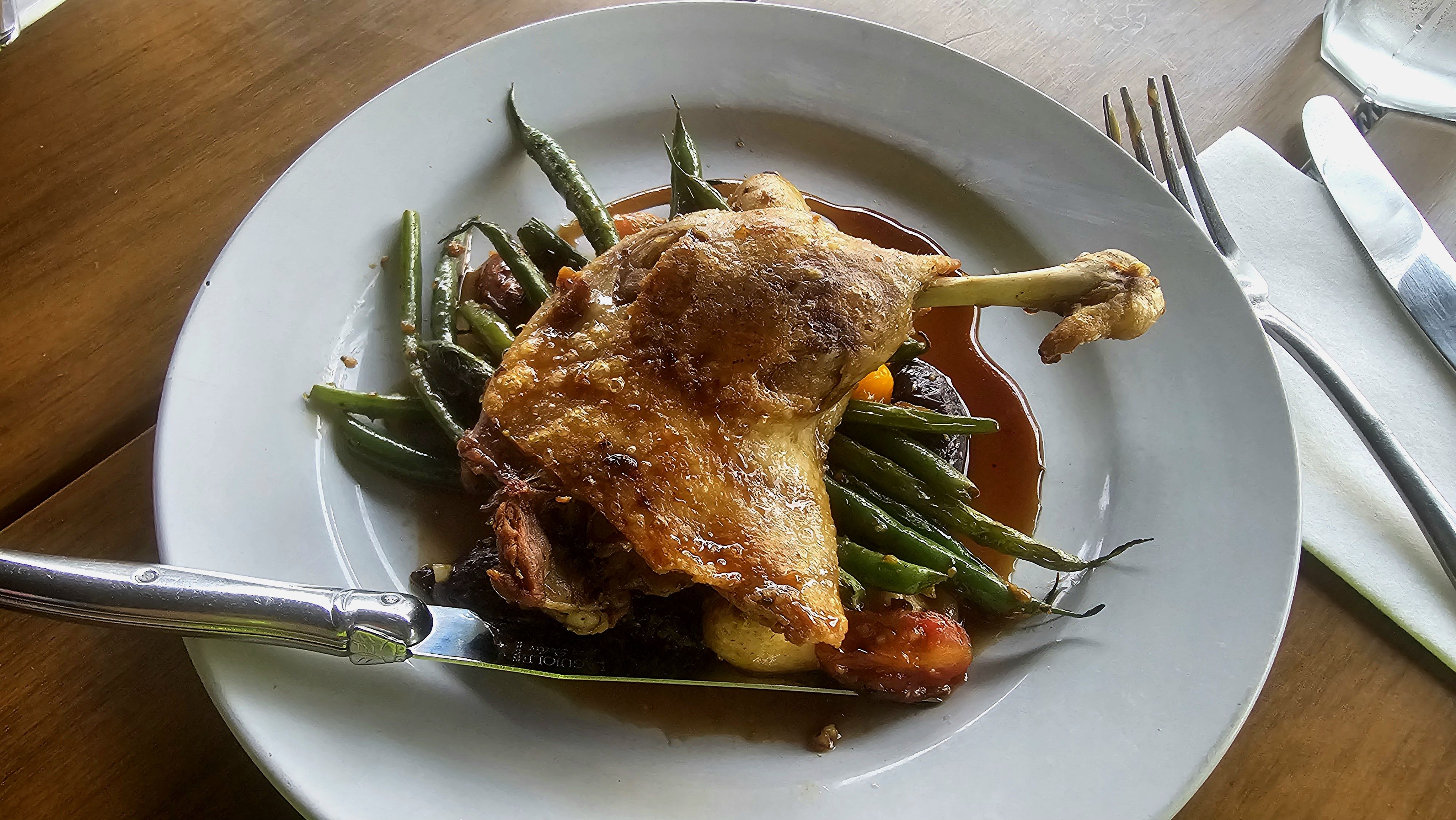 A roasted chicken leg sits on a bed of green beans with a brown sauce on a gray plate, set on a wooden table with a knife, fork, napkin, and a glass in the background.
