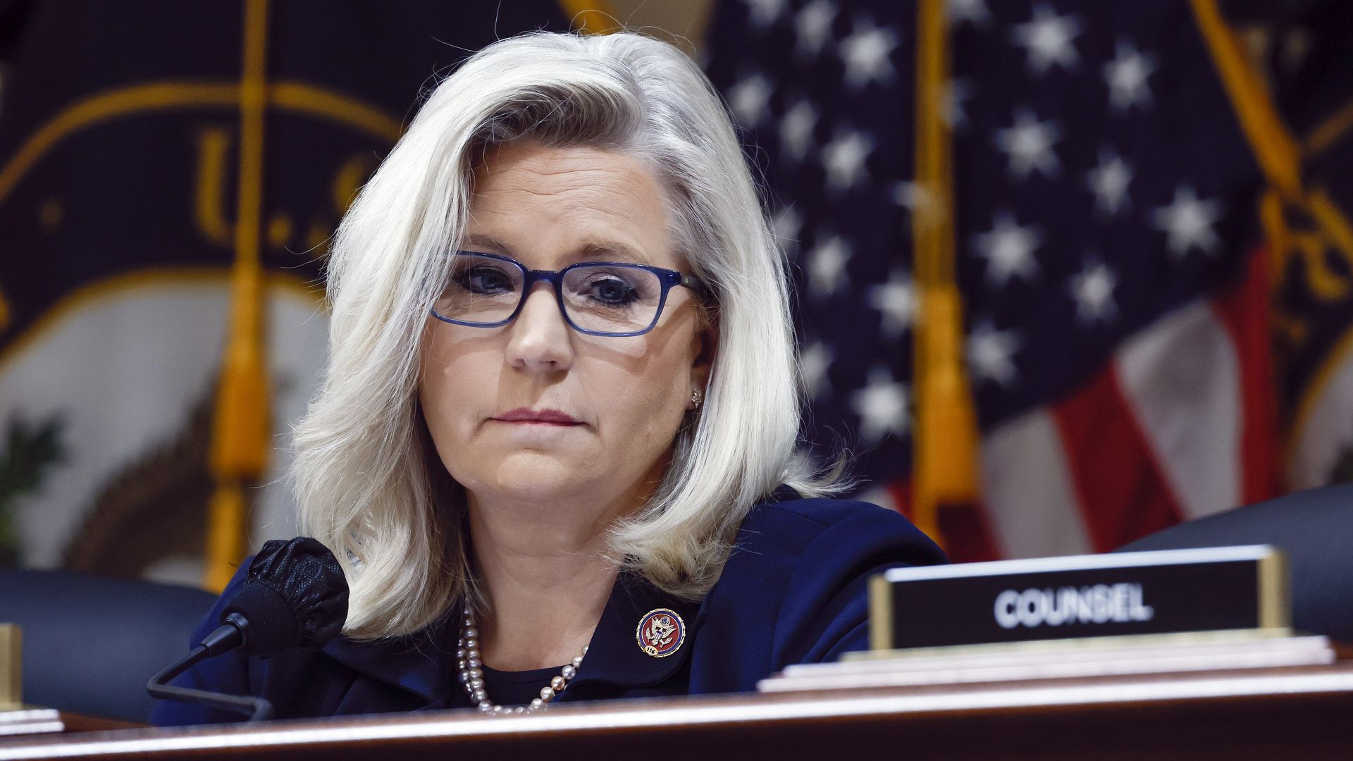 Rep. Liz Cheney, wearing a blue blazer, glasses and a pearl necklace, sits behind a podium at a Jan. 6 committee hearing.