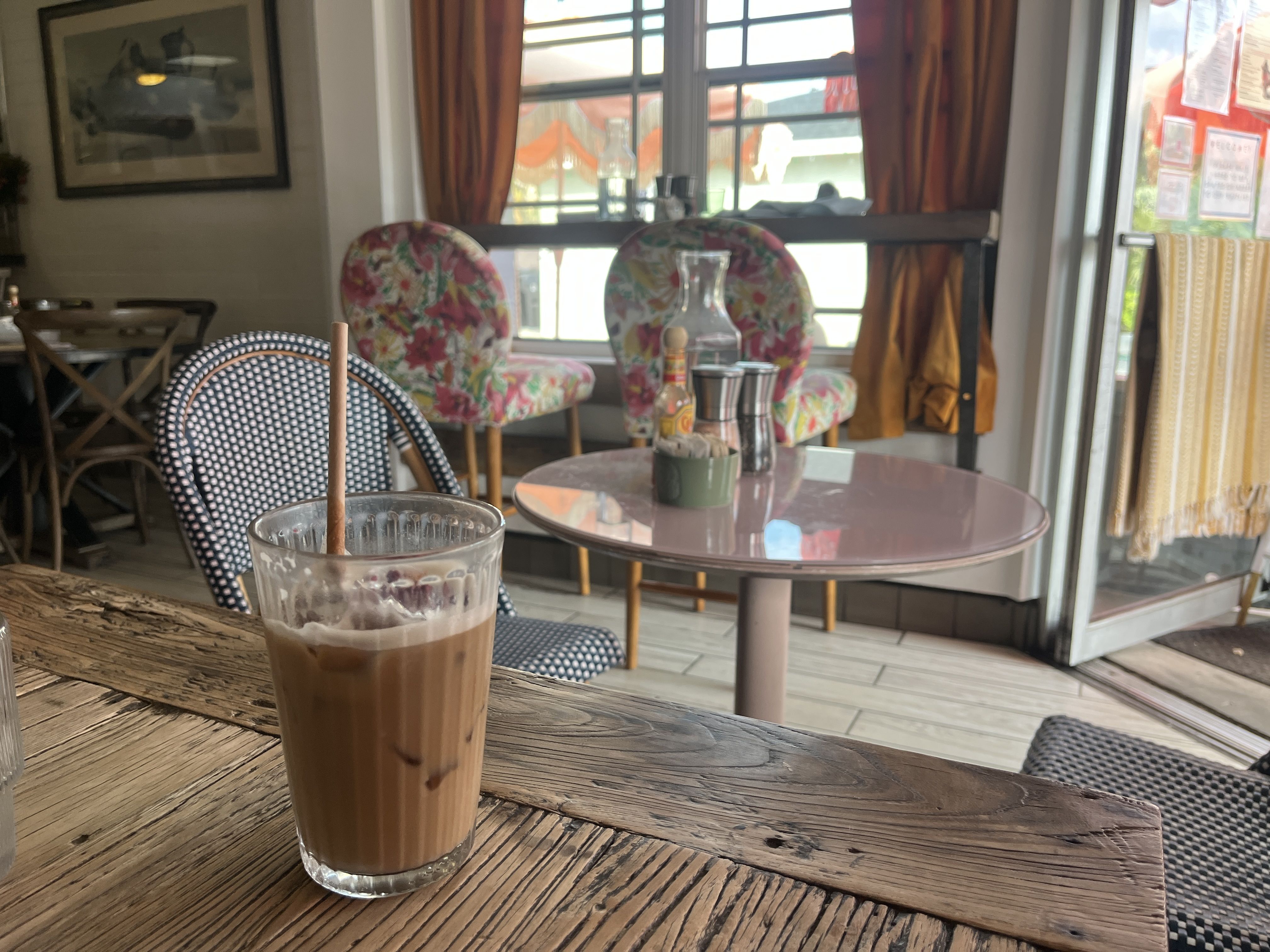 Glass of iced coffee with a brown straw on a rustic wooden table inside a cozy cafe with floral and patterned chairs, a round pink table, and a window with orange curtains.
