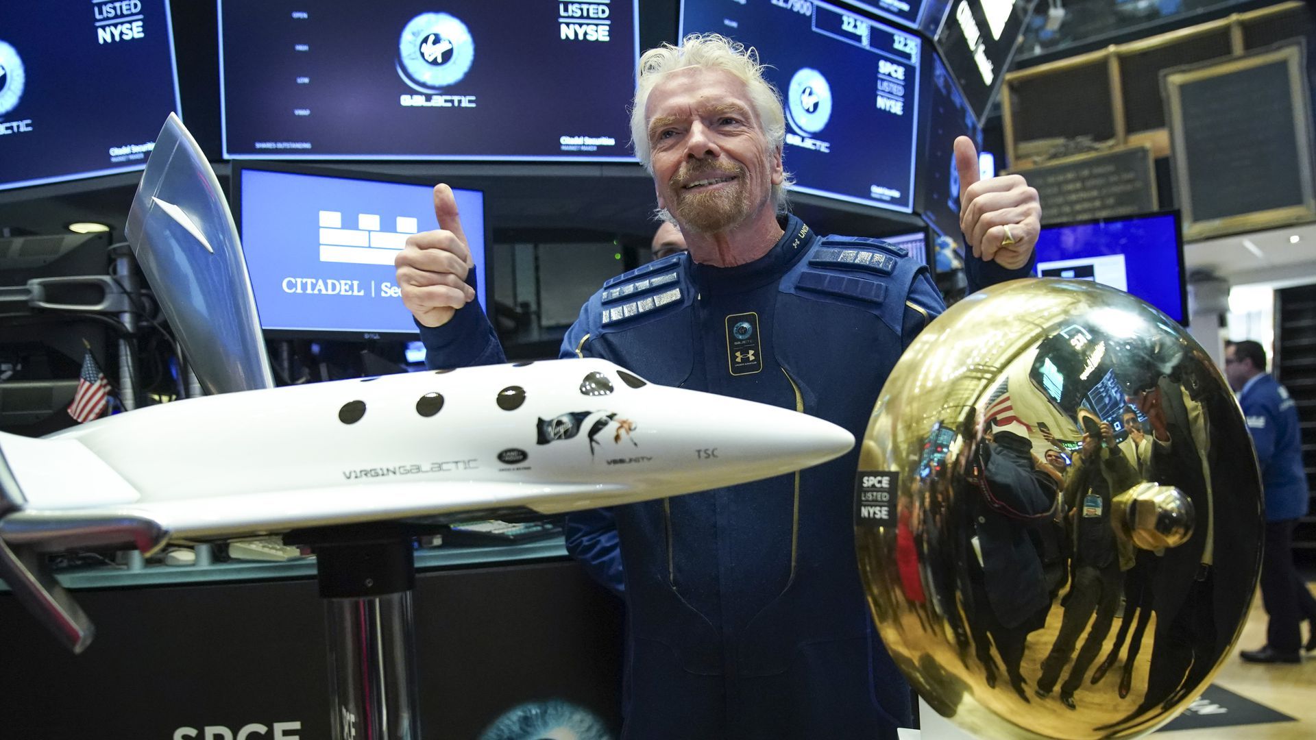 Photo of a white man with white hair holding both thumbs up behind a miniature model of an airplane 