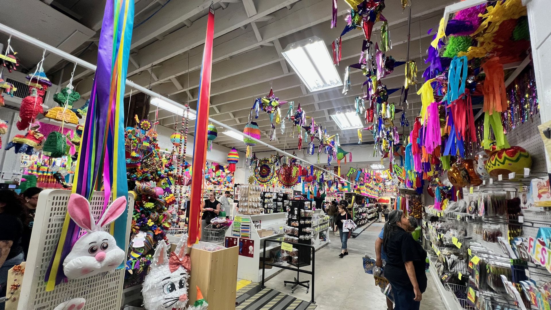 A busy scene at Amols' Party & Fiesta Favors in San Antonio shows a party supply store filled with vibrant, colorful decorations — ribbons, streamers, piñatas, and balloons — hanging from the ceiling while customers browse shelves and displays.