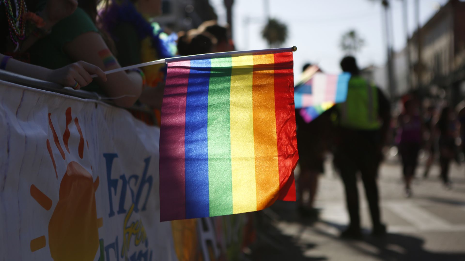 Rainbow Pride flag attached to a railing at a sunny street parade; vivid red, orange, yellow, green, blue, and purple stripes with blurred marchers in the background.