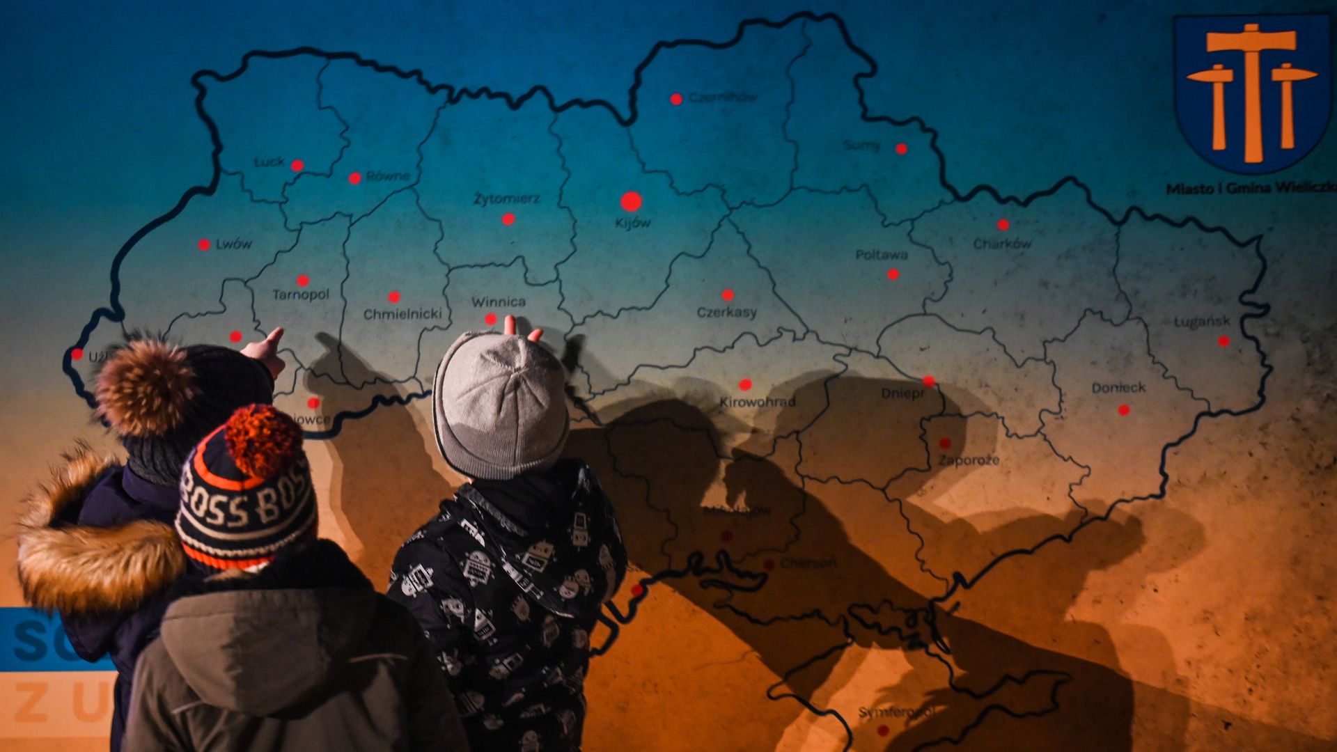 Young Ukrainian kids gathered around a board with a map of Ukraine, pointing to their hometowns with excitement during the 'Light for Ukraine' ceremony