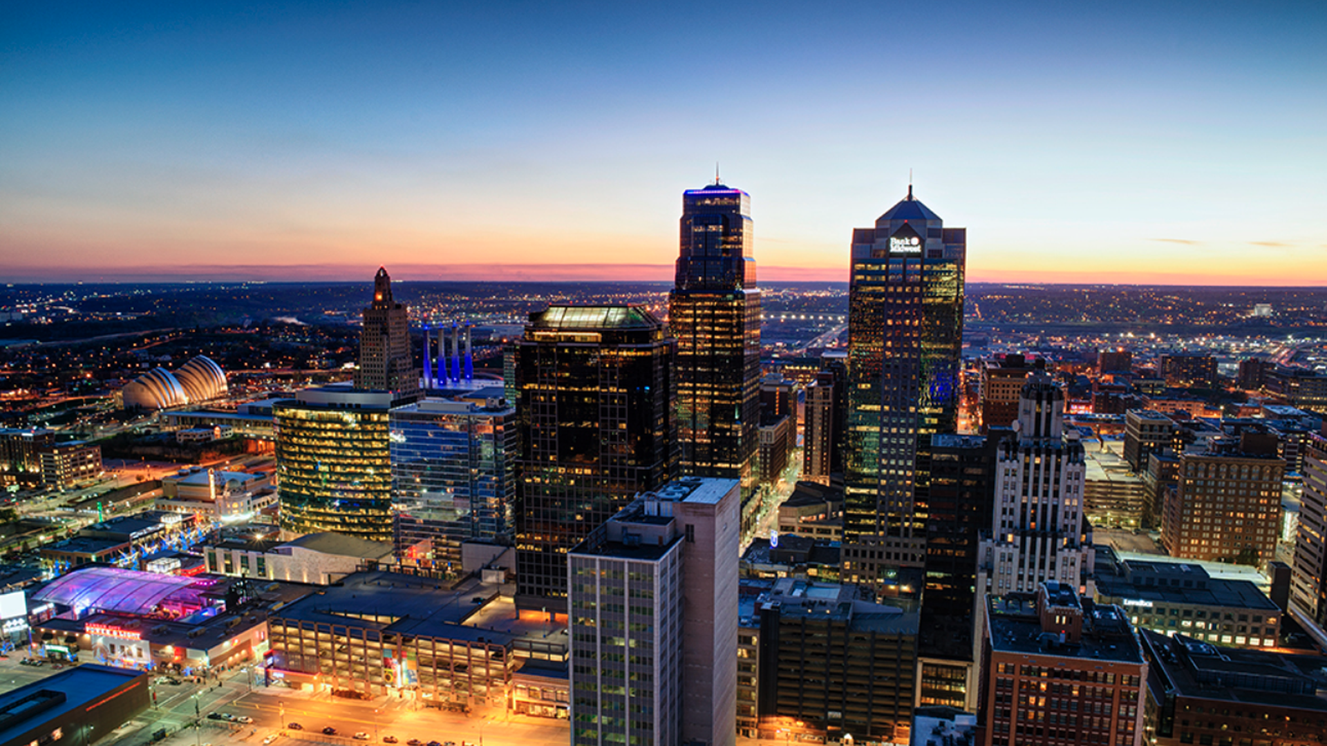 The Kansas City skyline at dusk with illuminated skyscrapers, including the Bank of America building, colorful lights on nearby structures, and a clear sky transitioning from orange to deep blue.