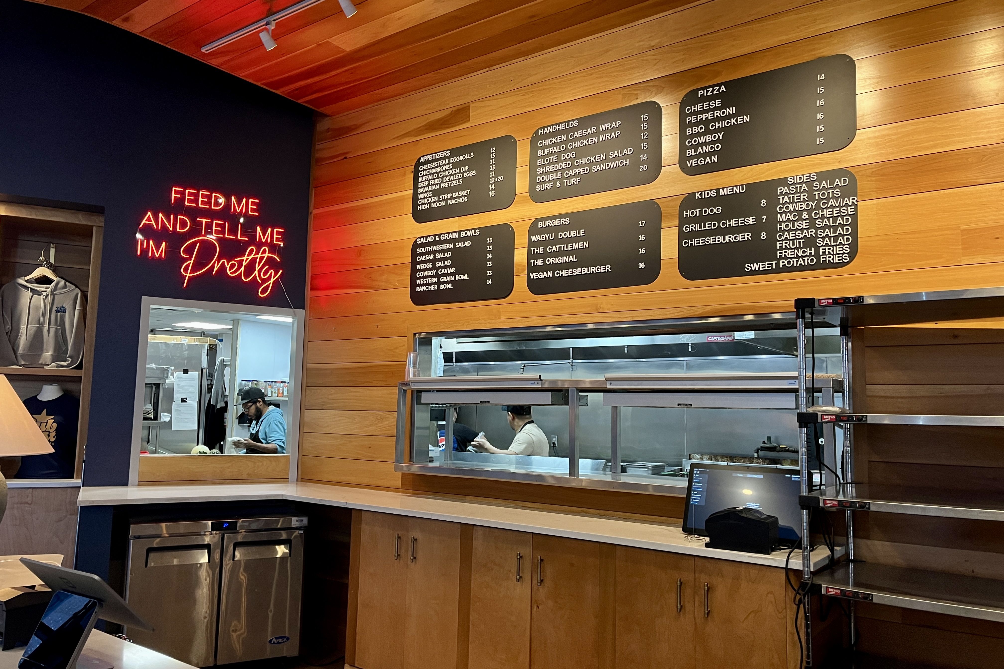Wood-paneled wall with black menu boards and a blue wall neon sign reading "FEED ME AND TELL ME I'M Pretty." An open kitchen window shows a worker; counter and shelves nearby.