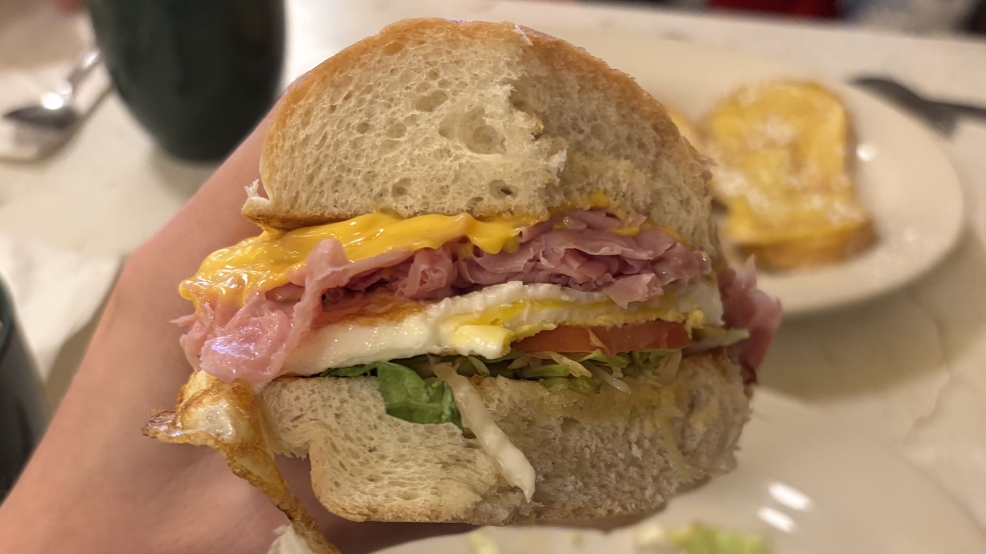 Hand holding a sandwich with white bread, melted cheddar cheese, ham, fried egg, lettuce, and tomato on a blurred plate with another sandwich in the background.