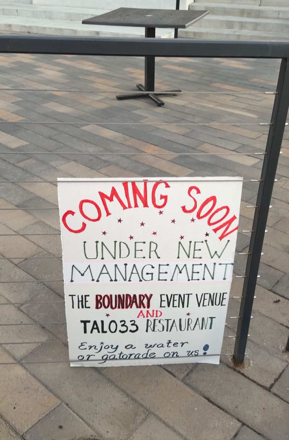 Outdoor plaza with a dark metal railing and a white poster reading "COMING SOON" in red and "UNDER NEW MANAGEMENT" in black; a small black table sits behind on the tiled ground.