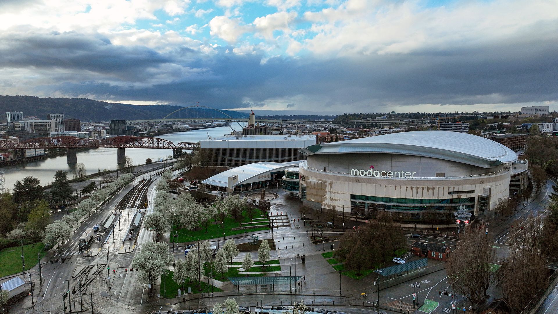Aerial cityscape featuring the circular Moda Center in the foreground, a river with two bridges, modern buildings, and tree-lined streets beneath a blue sky with dramatic clouds over distant hills.