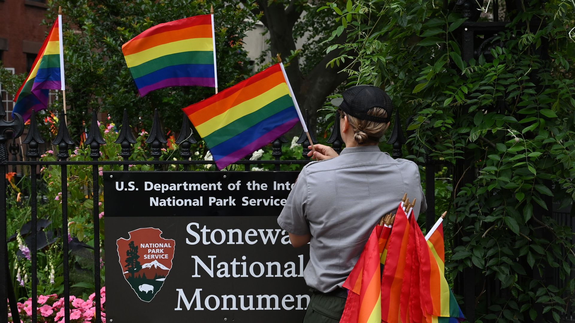 A person wearing a gray shirt and army green pants puts rainbow flags along a fence adorned with a sign that reads "U.S. Department of the Interior National Parks Service Stonewall National Monument."