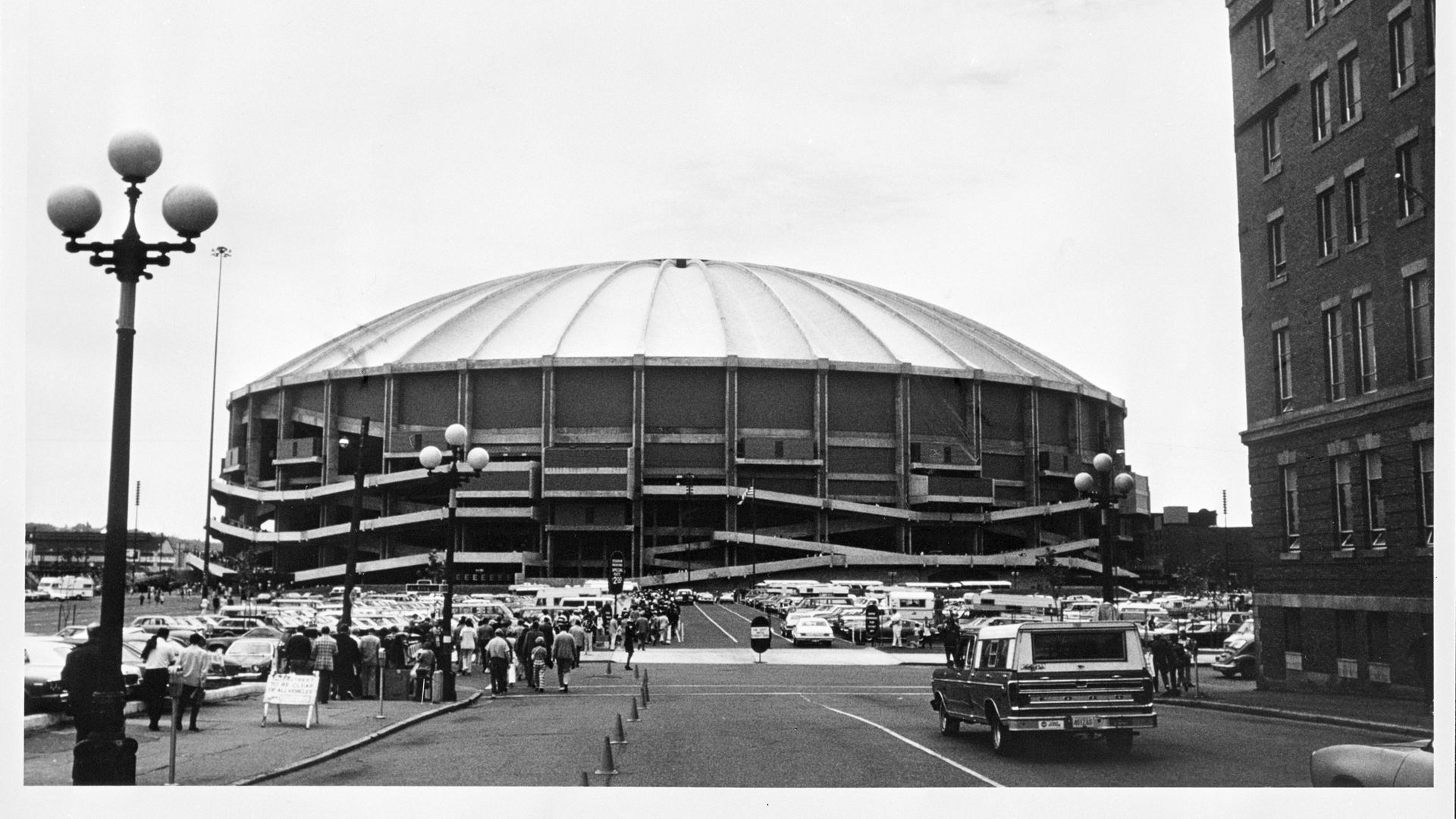 A black and white photo of the concrete Kingdome and crowds approaching it through a parking lot. An old fashioned lamp post is in the foreground along with a brick building. 