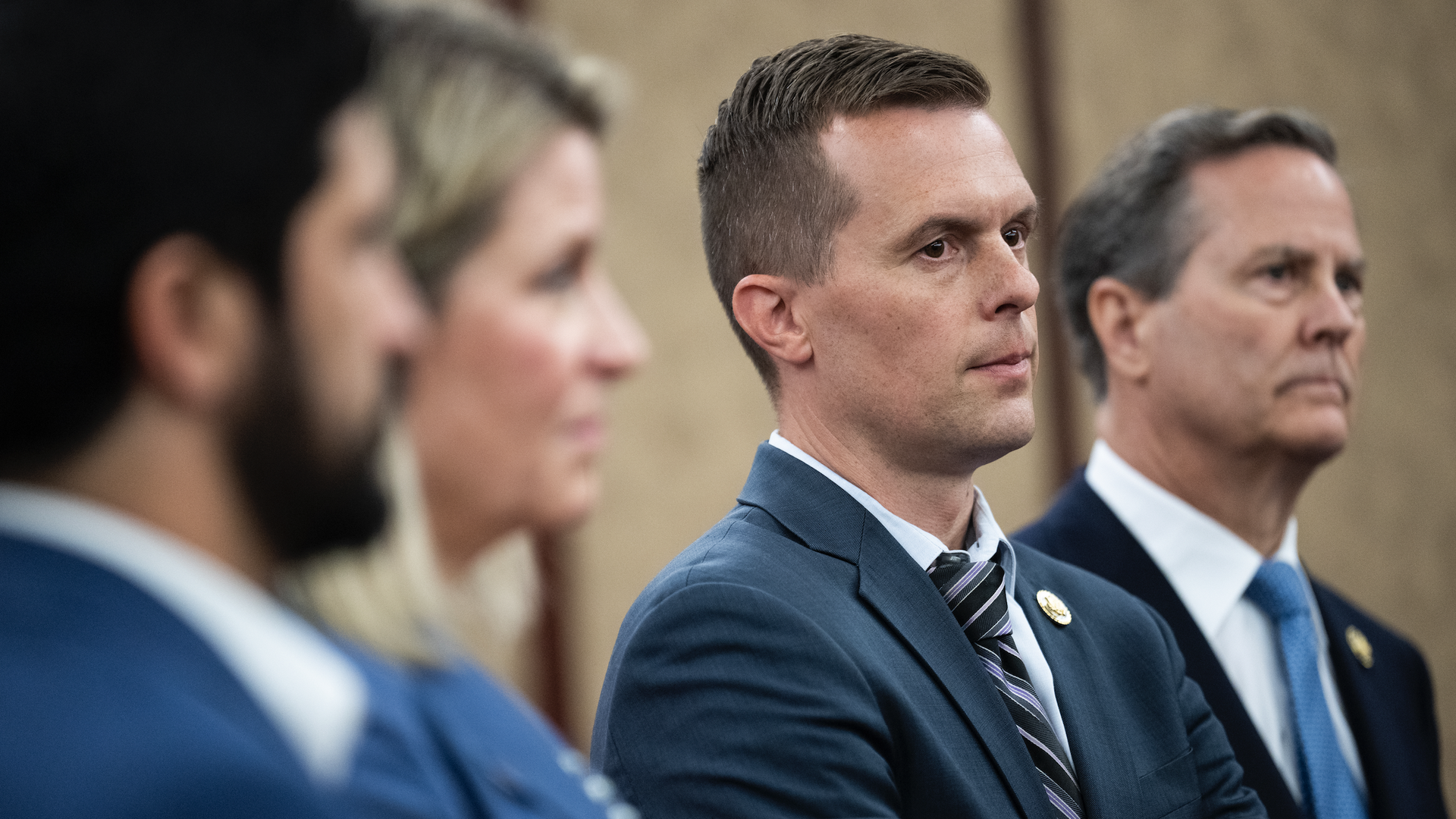 Rep. Jared Golden stands with colleagues while wearing a blue suit and standing in a beige room.