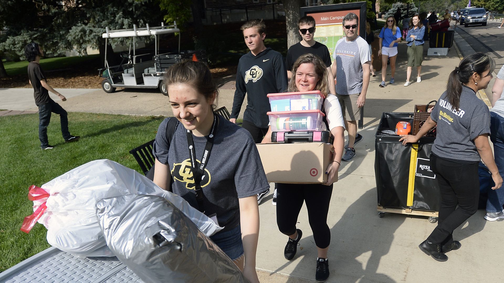 Students carrying boxes and bags while moving belongings on a sunny college campus sidewalk, some wearing gray shirts with a CU logo and a volunteer shirt, others walking nearby.