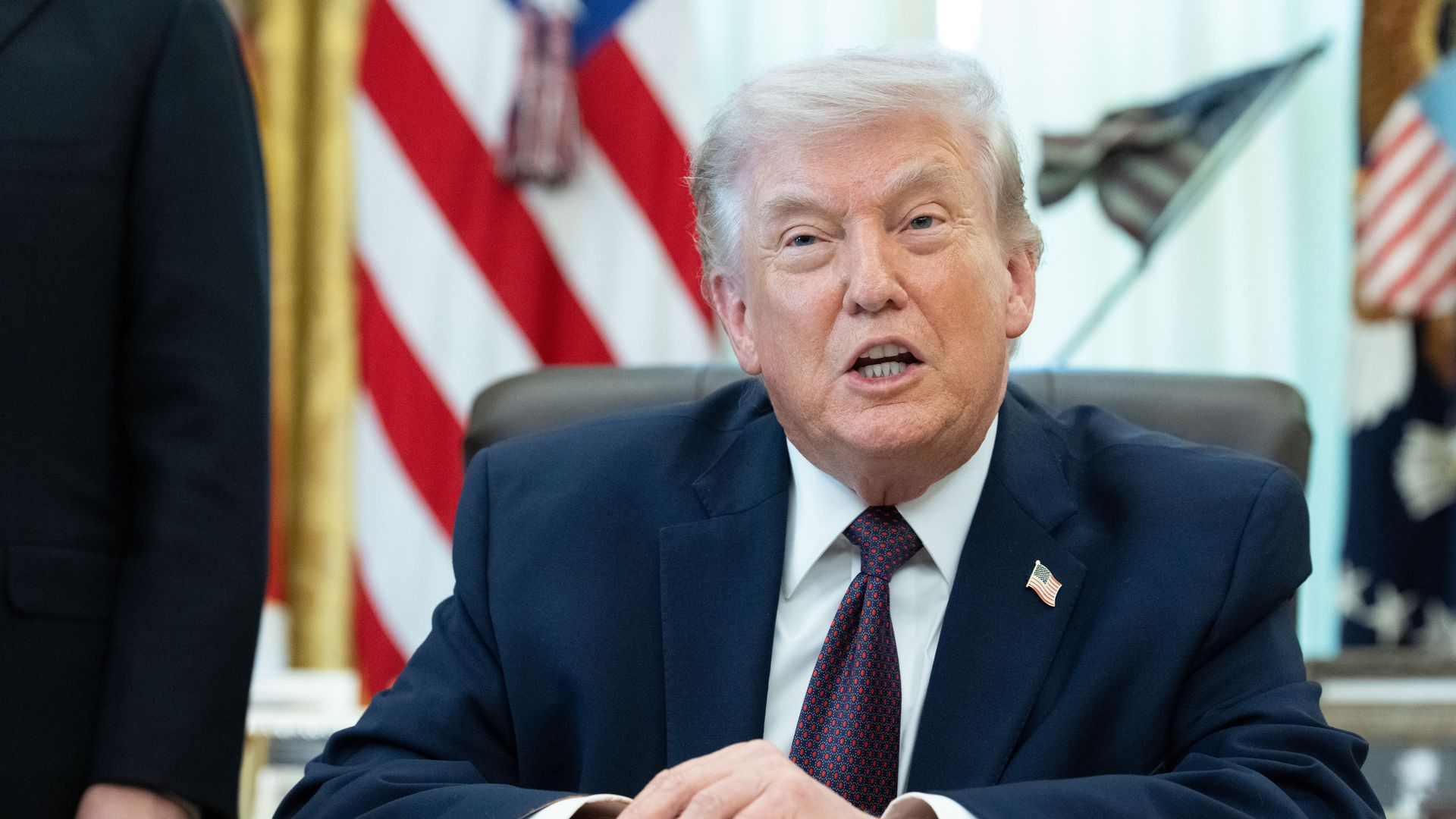 President Trump, with light hair in a dark blue suit and red tie sits at a desk, hands folded, speaking. Behind him are U.S. flags and office decor.
