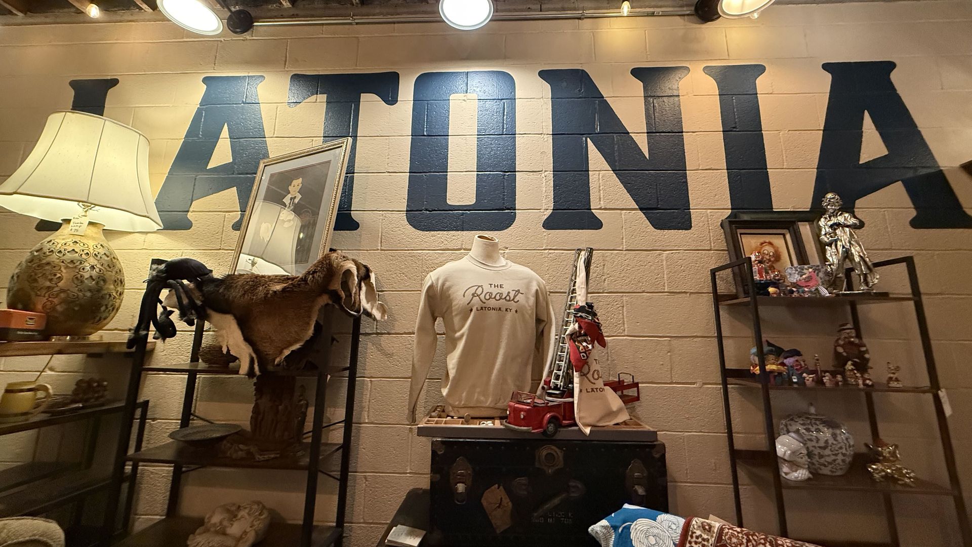 Vintage shop interior with a large blue "LATONIA" mural on a beige brick wall. Displays include a lamp, framed portrait, mannequins in tees, fur hides, and glass shelves of trinkets.