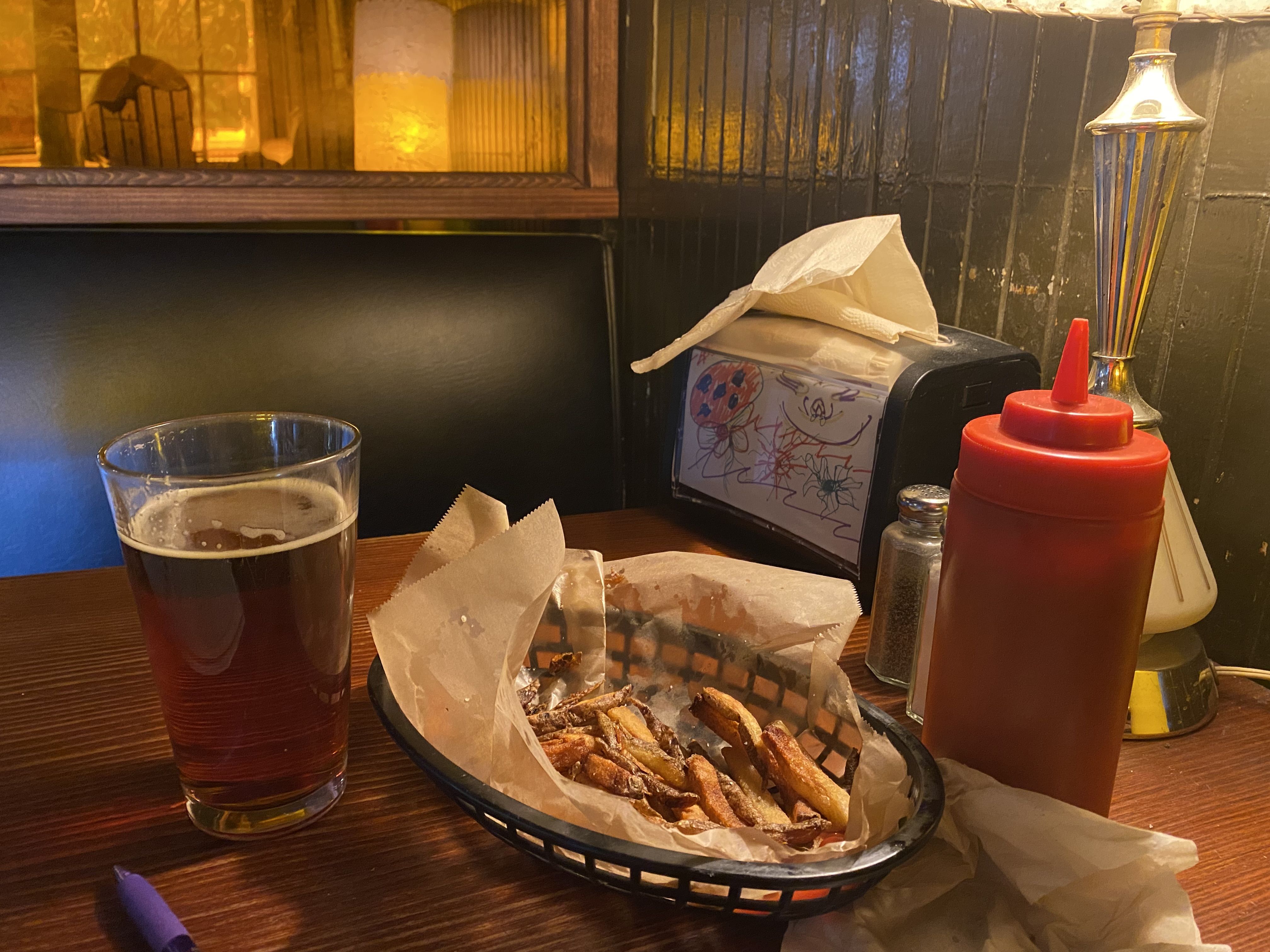 An almost empty basket of fries next to a squeeze bottle of ketchup on a booth table with green walls and a napkin holder.