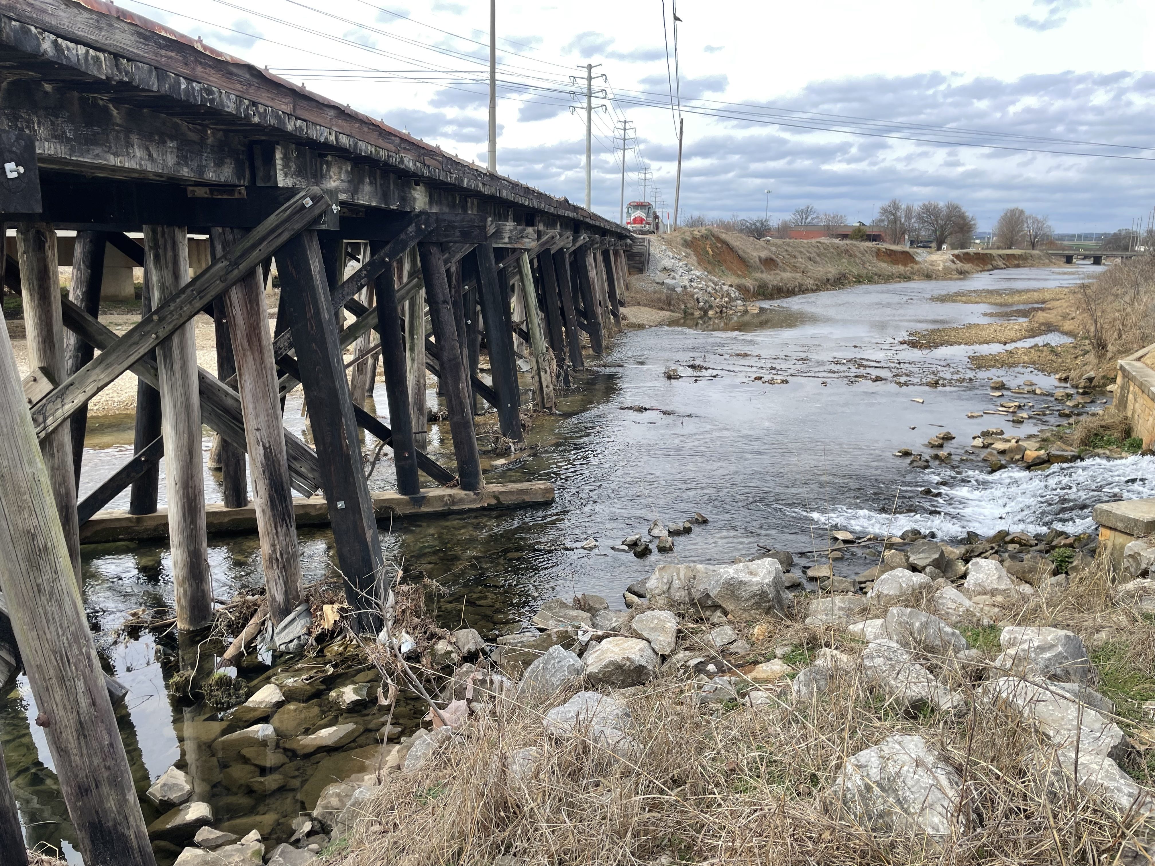 Wooden train trestle bridge over a rocky river with dry grasses and cloudy sky, a train approaching in the distance.