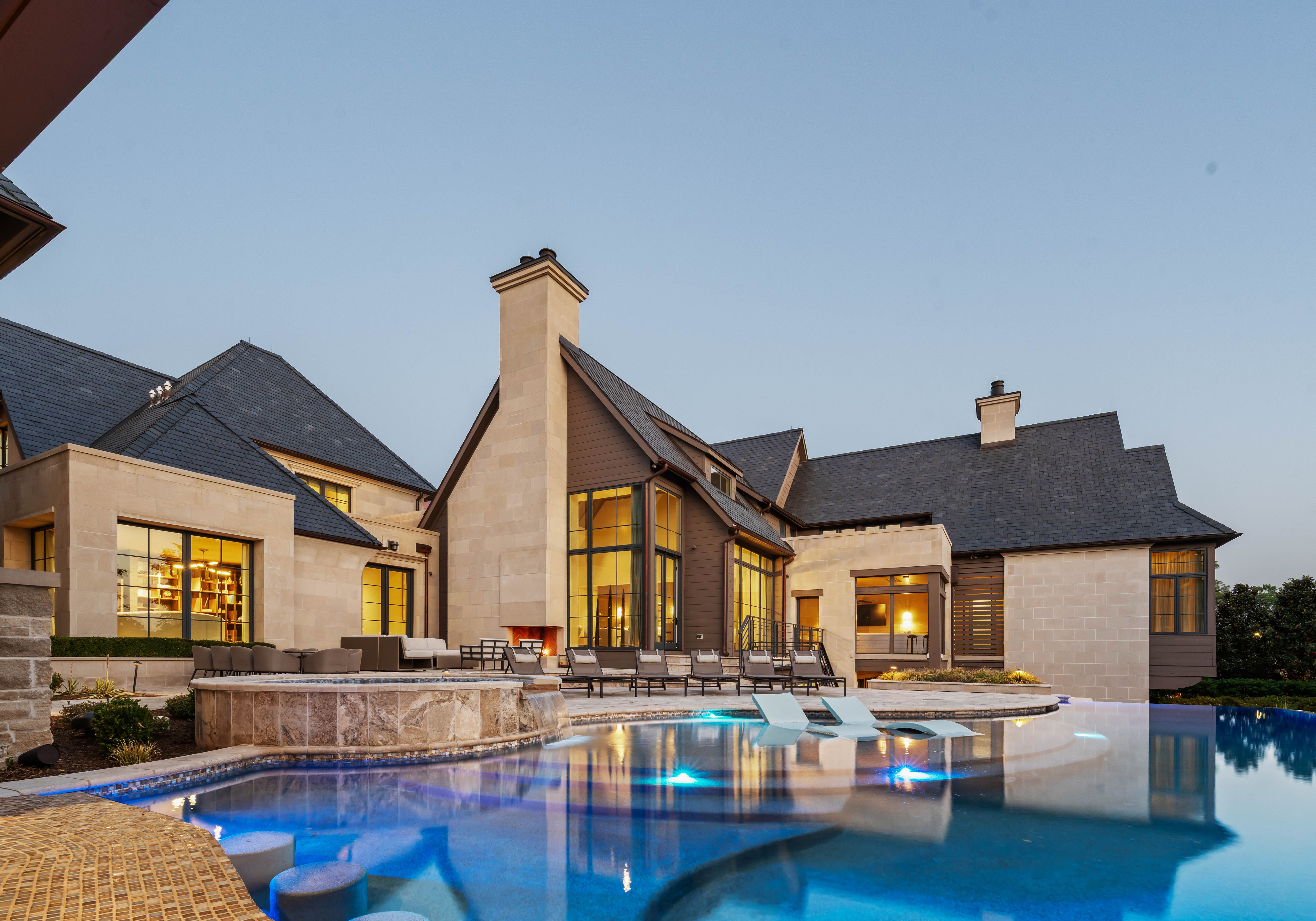 A modern beige stone house with slate roofs and large glass windows overlooks a lit pool. Outdoor seating lines the pool deck as dusk settles, with chimneys and warm interior lights.