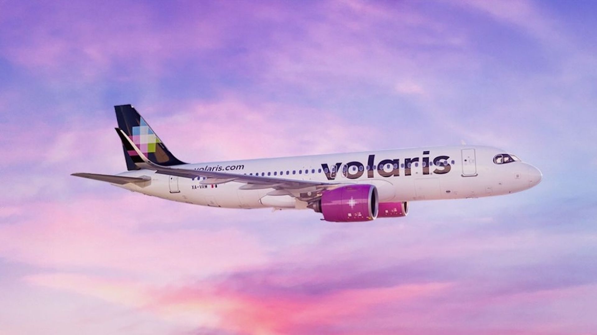 A white Volaris airliner flies left to right against a pink and purple sunset sky; large Volaris branding on the fuselage, purple engine nacelles, and a colorful patterned tail.