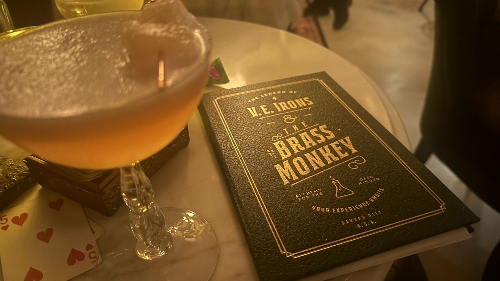 Close-up of a cocktail glass with an amber drink and frothy top on a marble table next to a black leather menu with gold lettering reading V.E. Irons & The Brass Monkey.