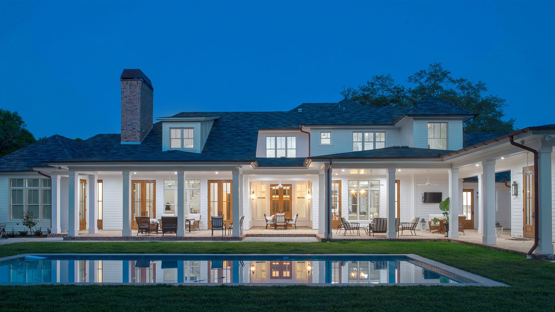 A house with wood doors, white beams and an open patio is reflected in a pool in the evening