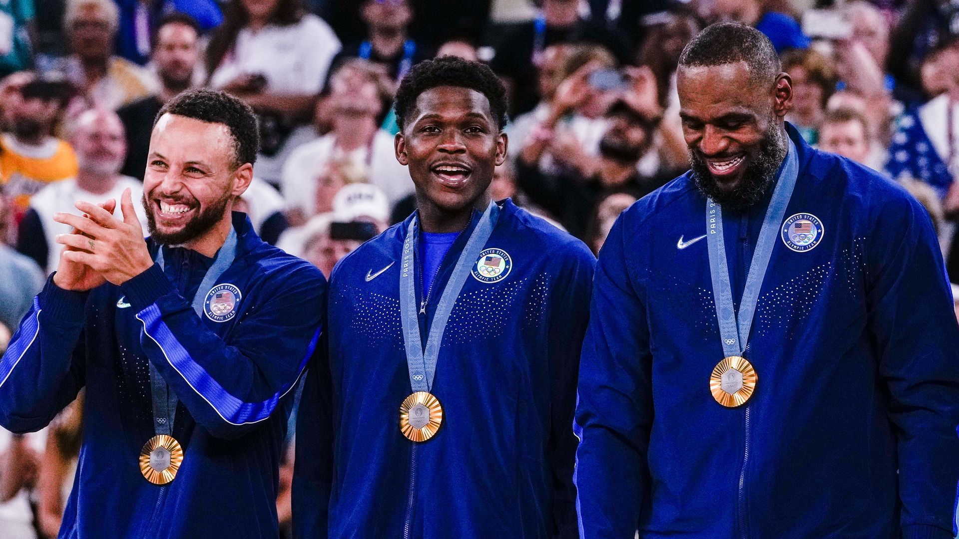 Three USA basketball players, Stephen Curry, Anthony Edwards, and LeBron James, smile while wearing blue tracksuits and Olympic gold medals around their necks