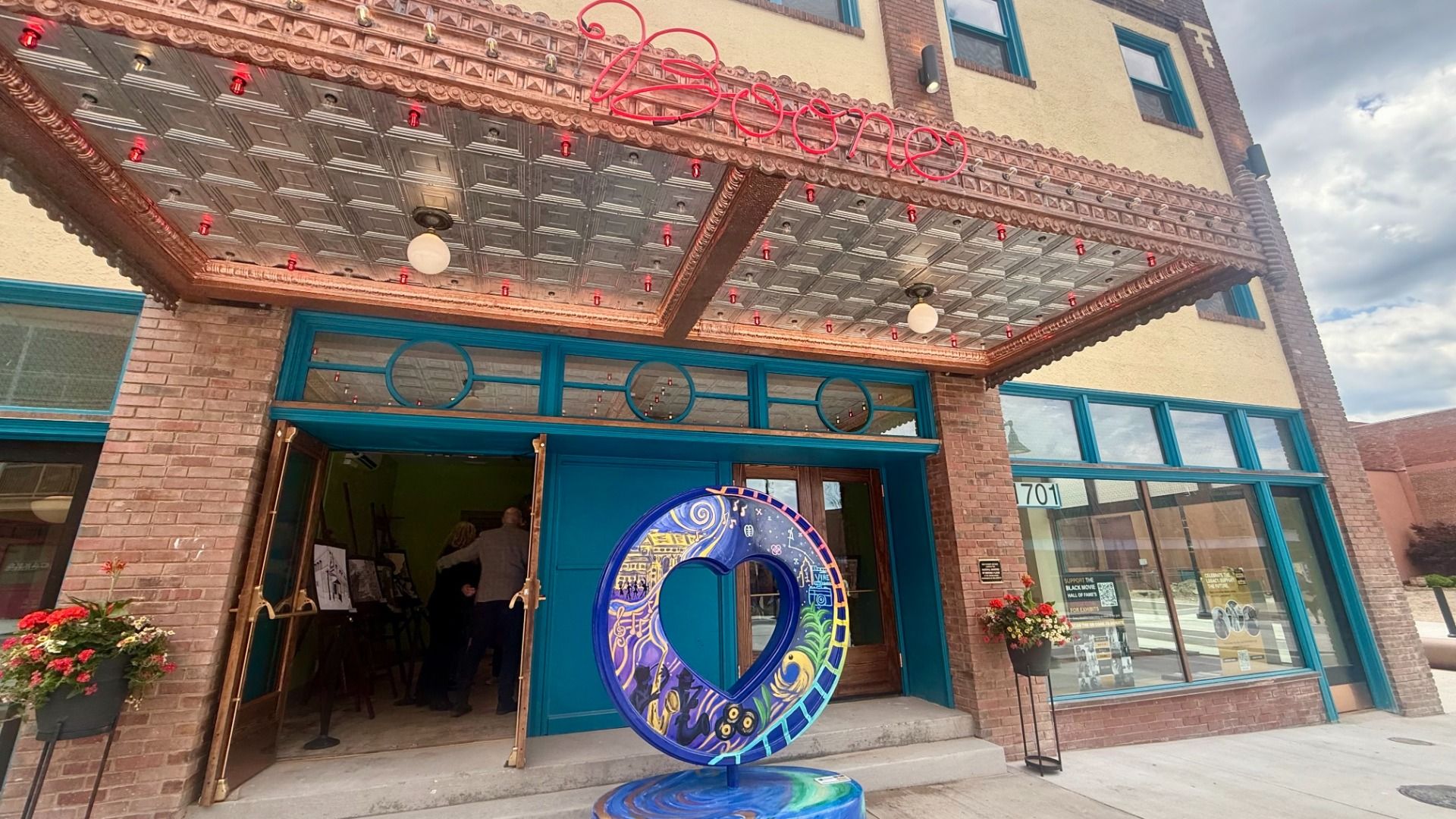 Teal-framed brick storefront with a blue doorway. A tin canopy with red lights extends over the entrance. A colorful circular sculpture with a heart opening sits outside, next to potted flowers.