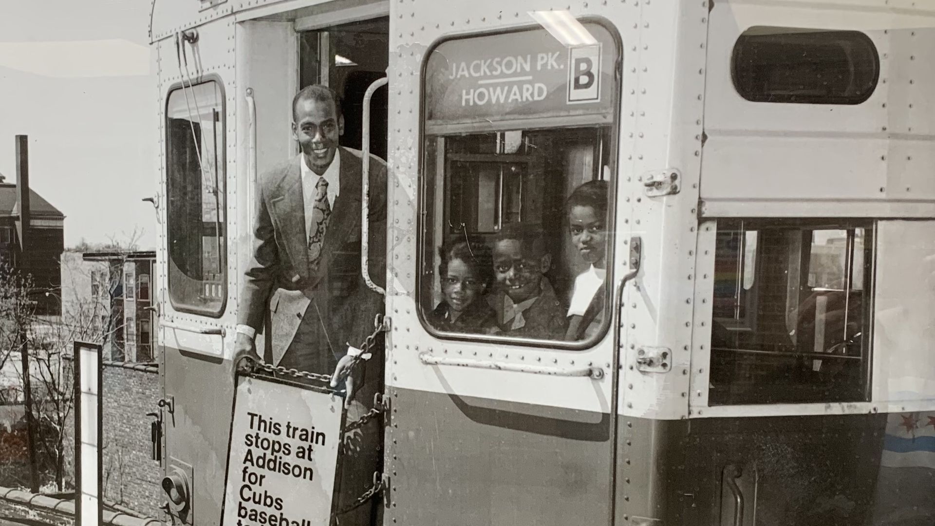 Black and white photo of a man in a suit and three children on a train labeled "Jackson Pk. Howard B" with a sign saying, "This train stops at Addison for Cubs baseball today."