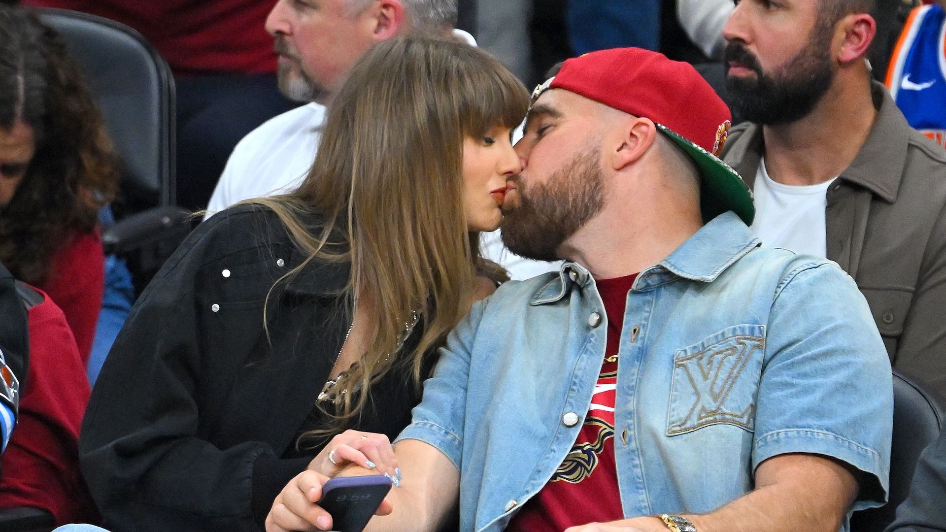 Couple kissing in a crowded stadium: a bearded man in a red cap and denim jacket leans toward a woman with long brown hair wearing a black jacket.