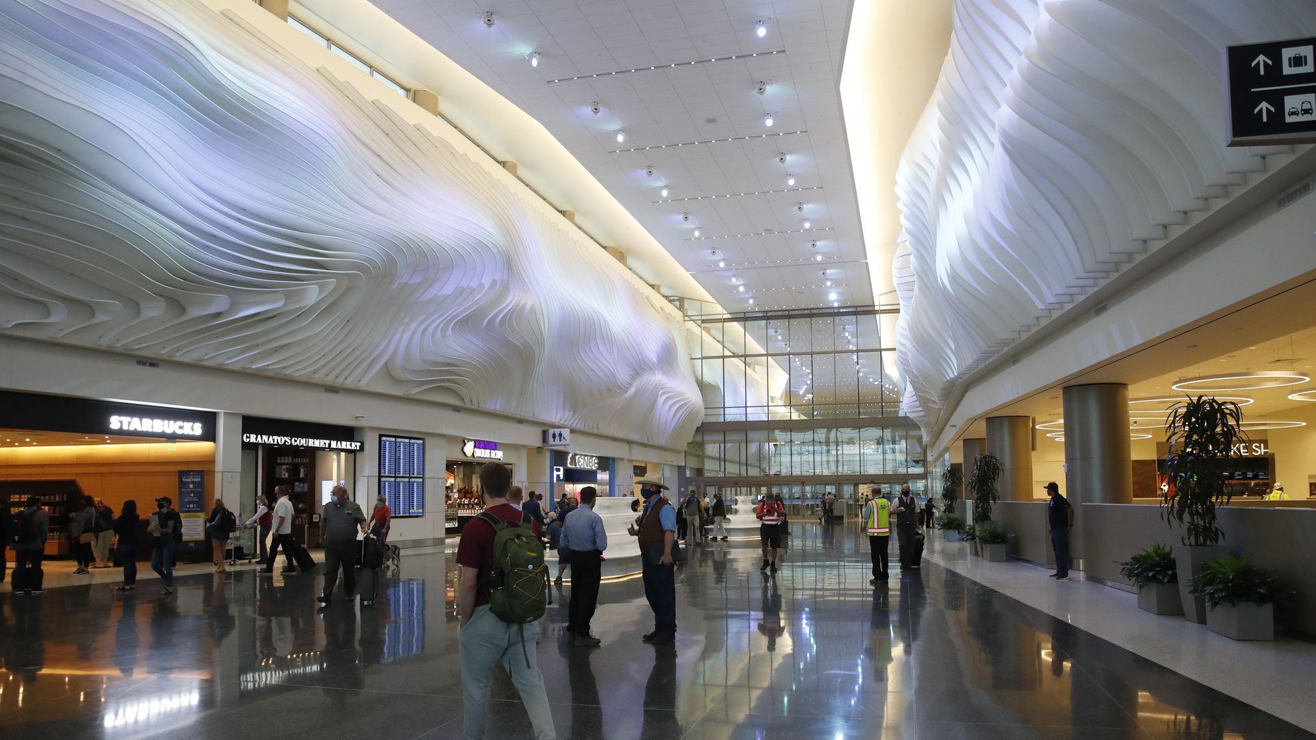 Bright modern airport terminal with white wavy ceiling design, reflective polished floor, shops like Starbucks, and travelers walking or standing with bags.