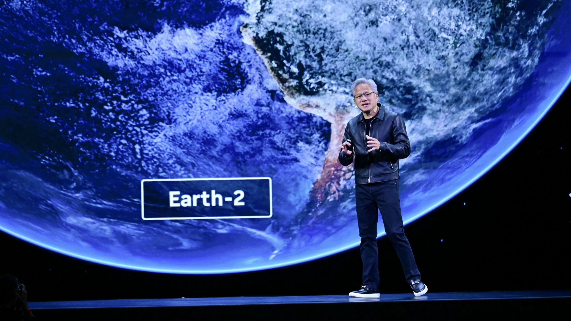 A person in a black leather jacket and sneakers speaking on stage in front of a large screen showing a blue and white Earth labeled "Earth-2".