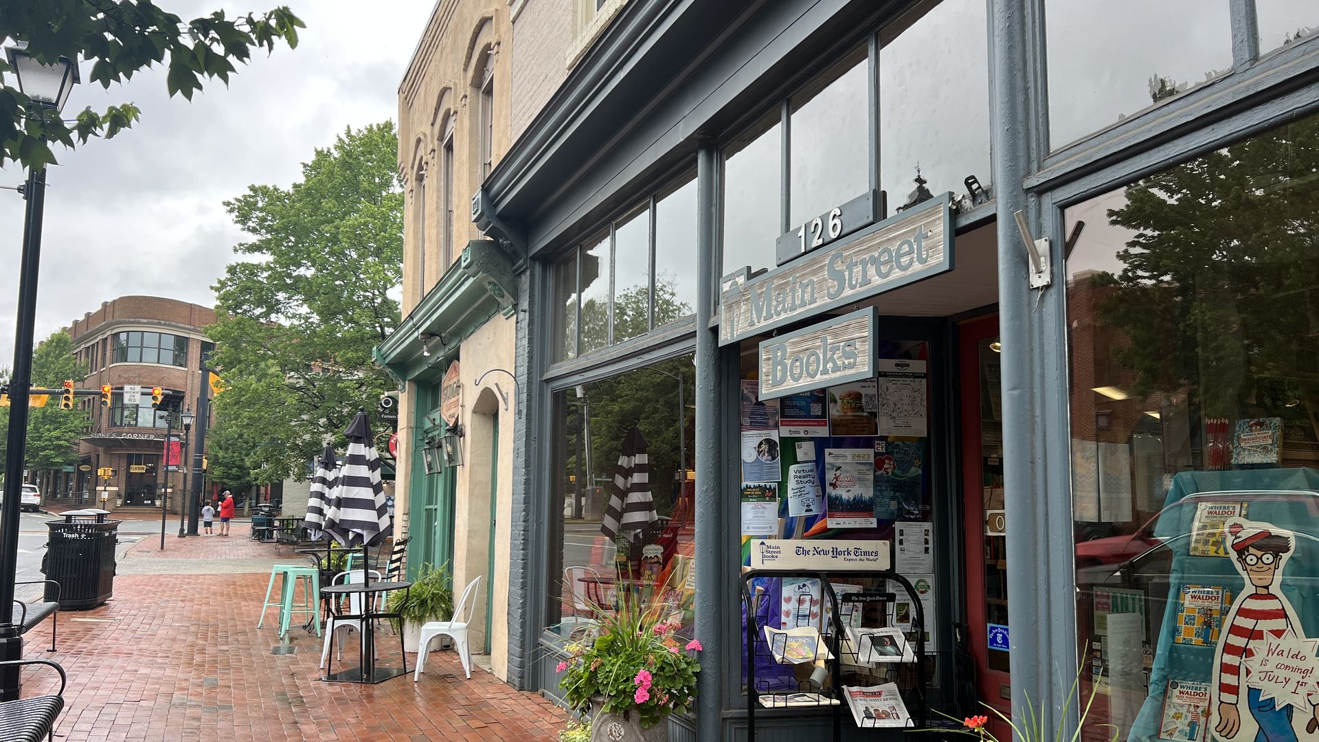 Brick sidewalk street view showing Main Street Books storefront with display racks, plants, and a Waldo poster, striped umbrellas, chairs, and buildings under cloudy sky.