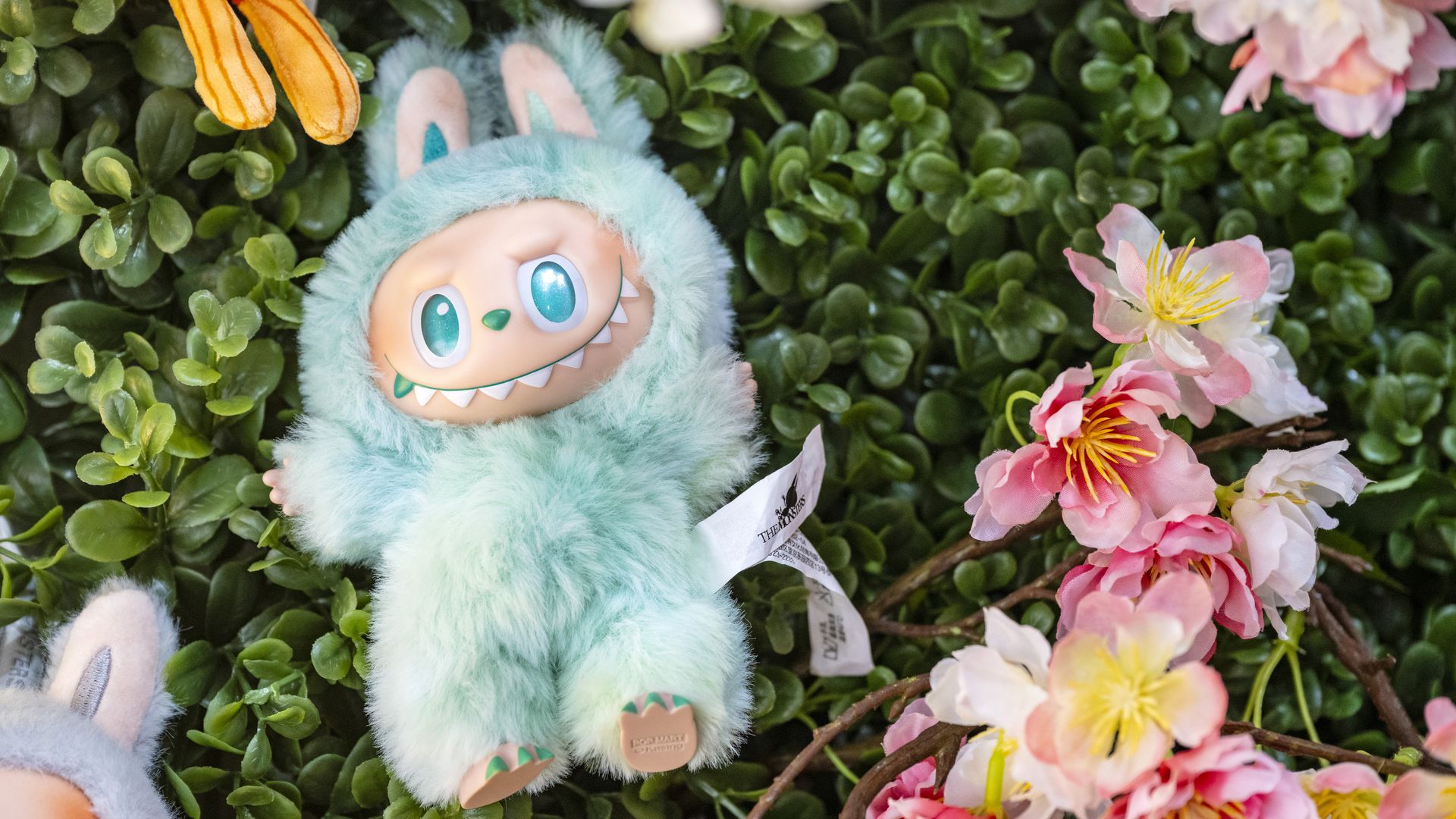 A Labubu doll on display on what looks like leaves or grass with flowers next to it