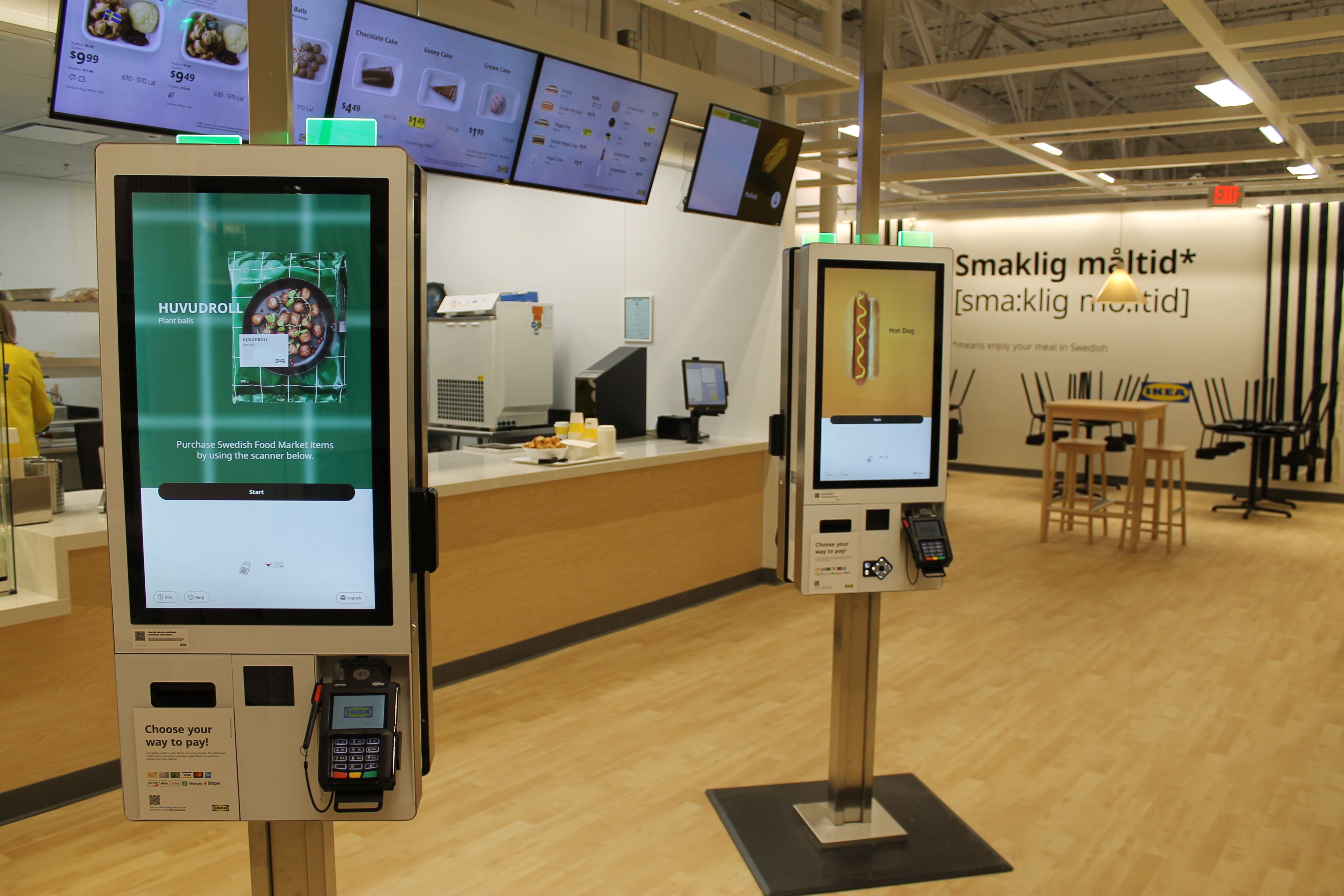 Two self-service kiosks with touchscreens and card readers in a light wood-floored IKEA food market area. Screens show plant balls and hot dog options. Tables and signage in background.