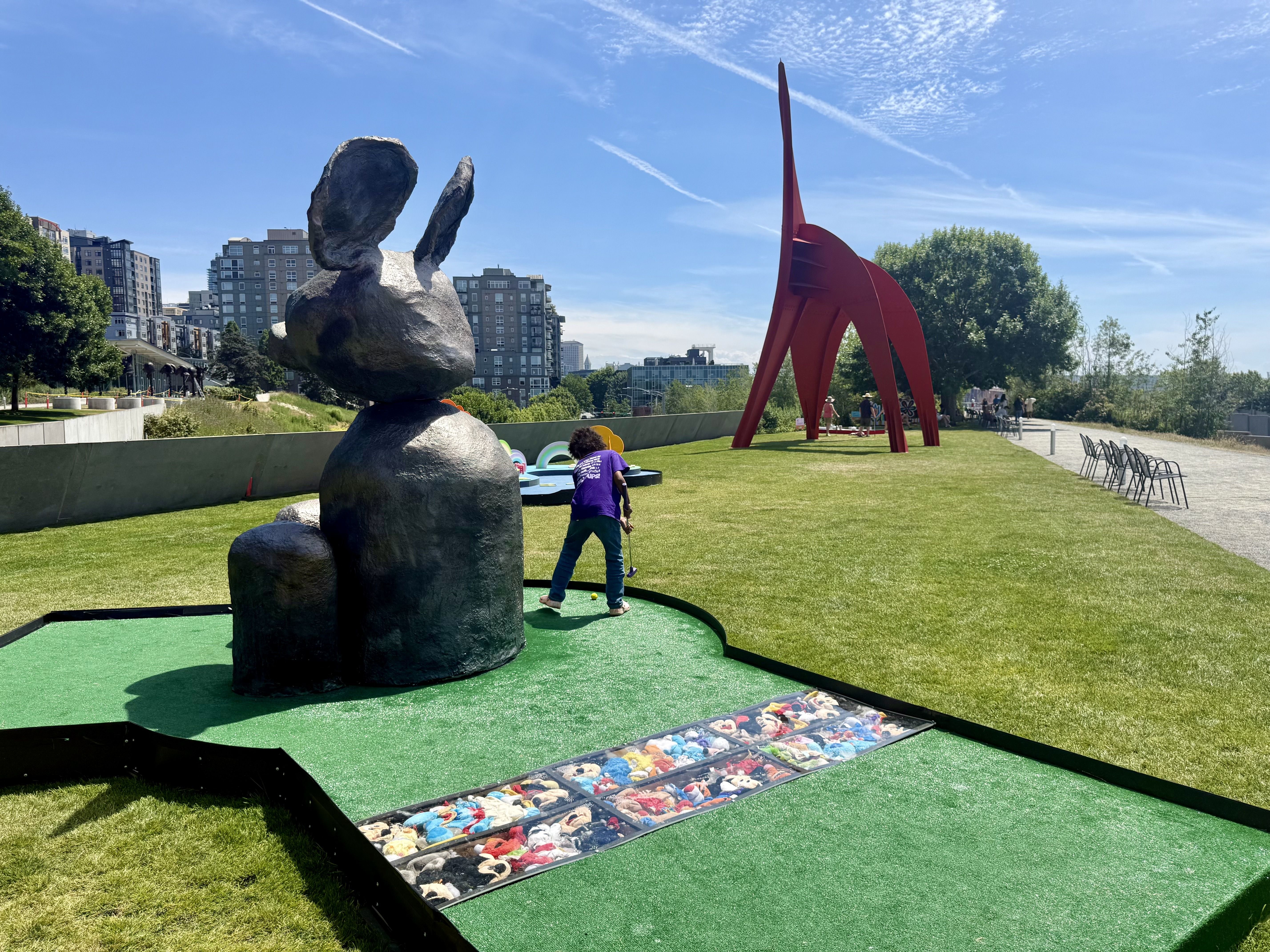 Sunny urban park with a large stacked-rock sculpture and minigolf station at left, and a tall red arched sculpture right. A child in purple plays mini-golf on green turf, with modern buildings, trees, and a blue sky.