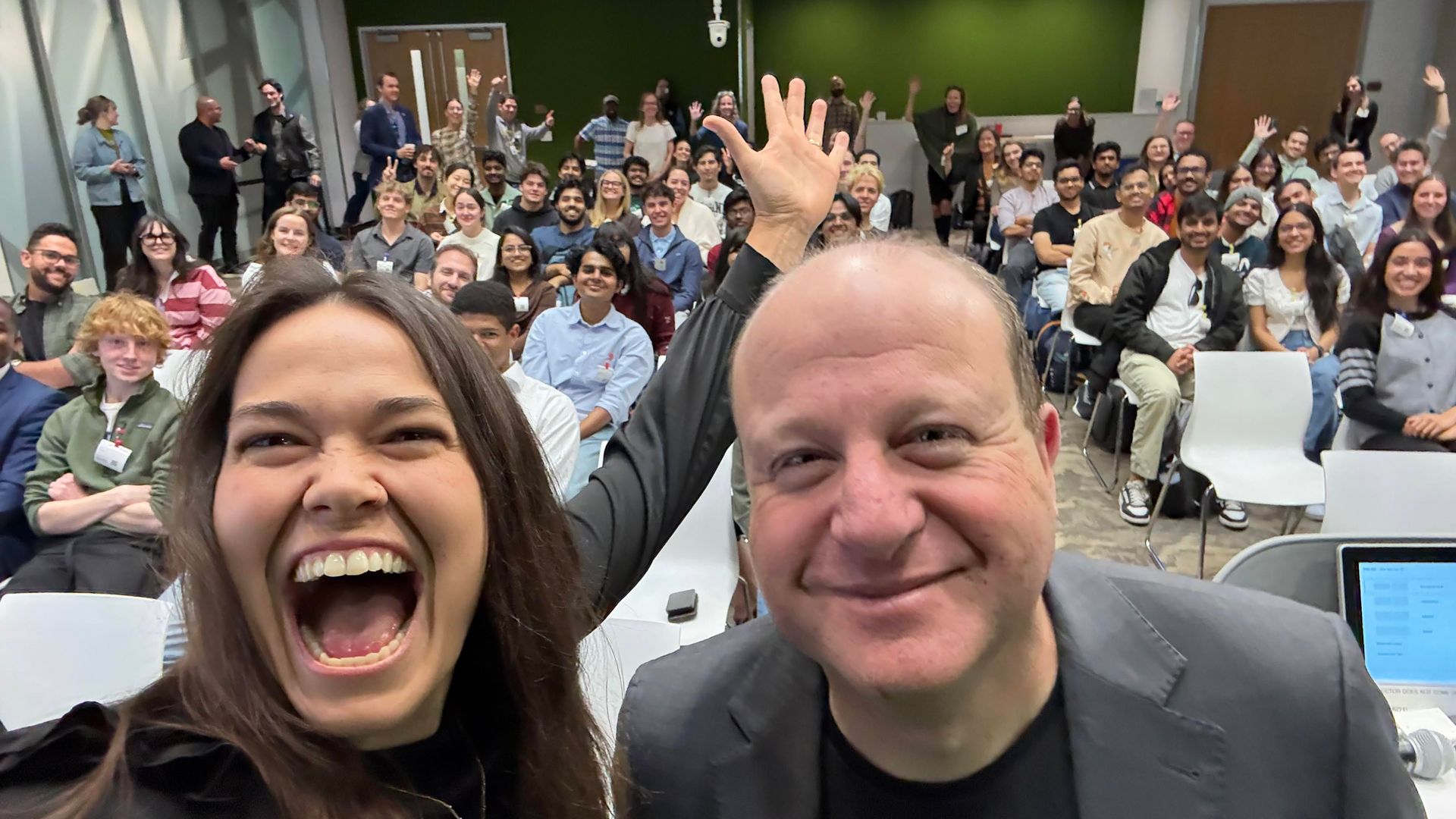 A large diverse group of people sitting in a conference room smiling and waving, with Google host Brin Enterkin and Colorado Gov. Jared Polis smiling close to the camera at the front.